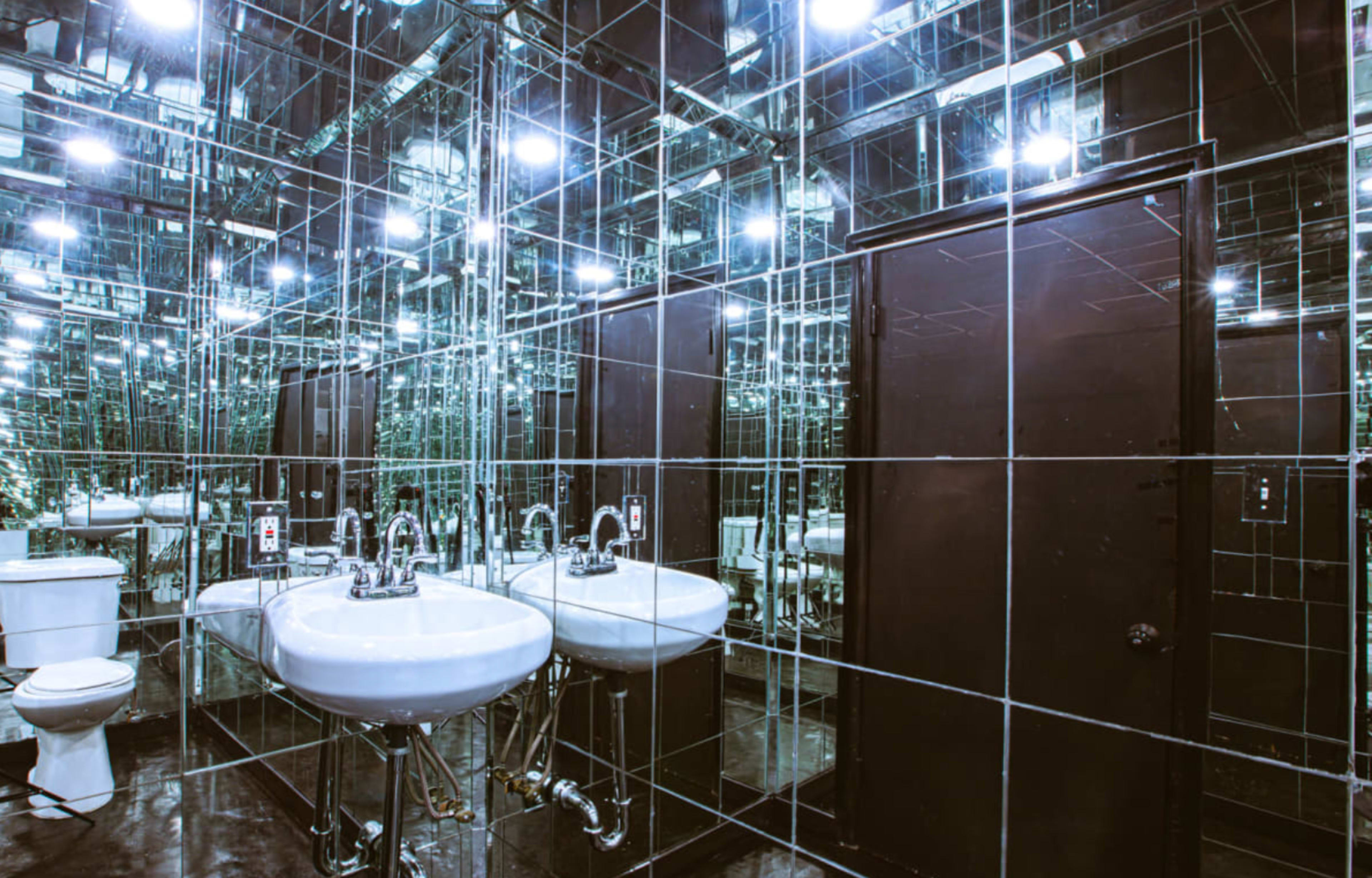 A modern restroom features mirrored walls, two sinks, a toilet, and bright overhead lighting.