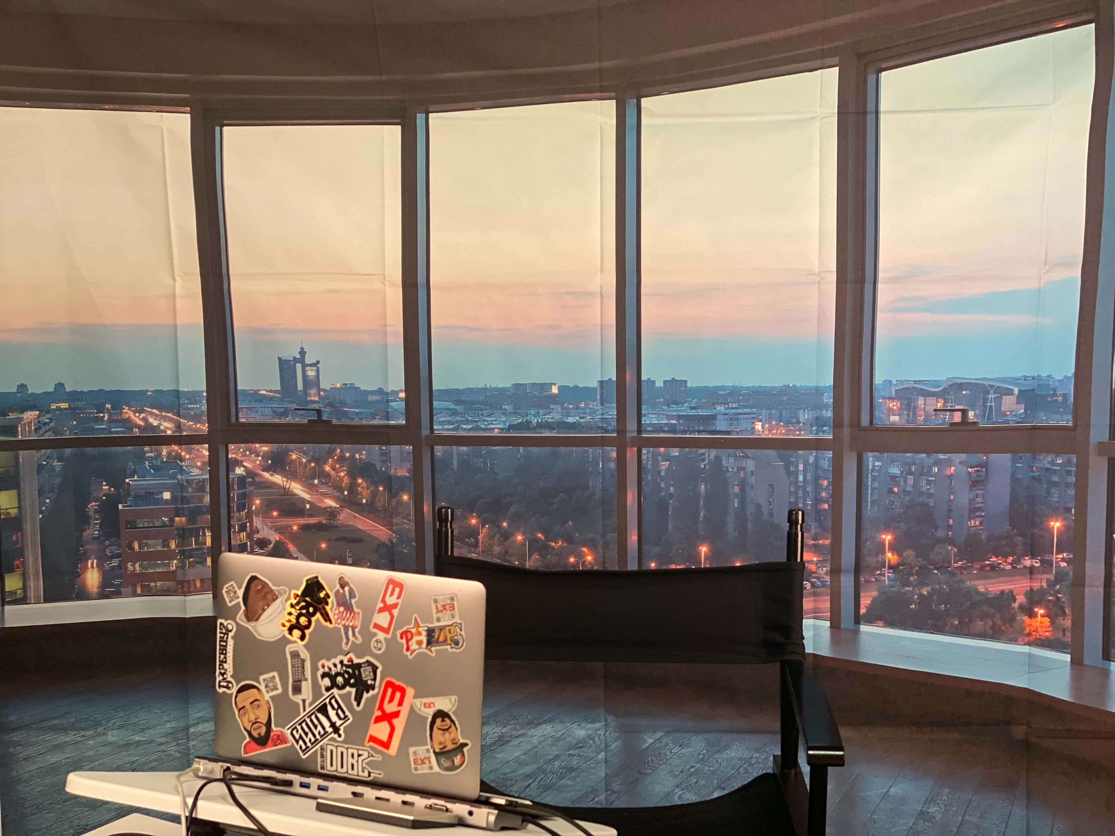 A laptop with stickers sits on a desk in front of large windows showcasing a city skyline at sunset.