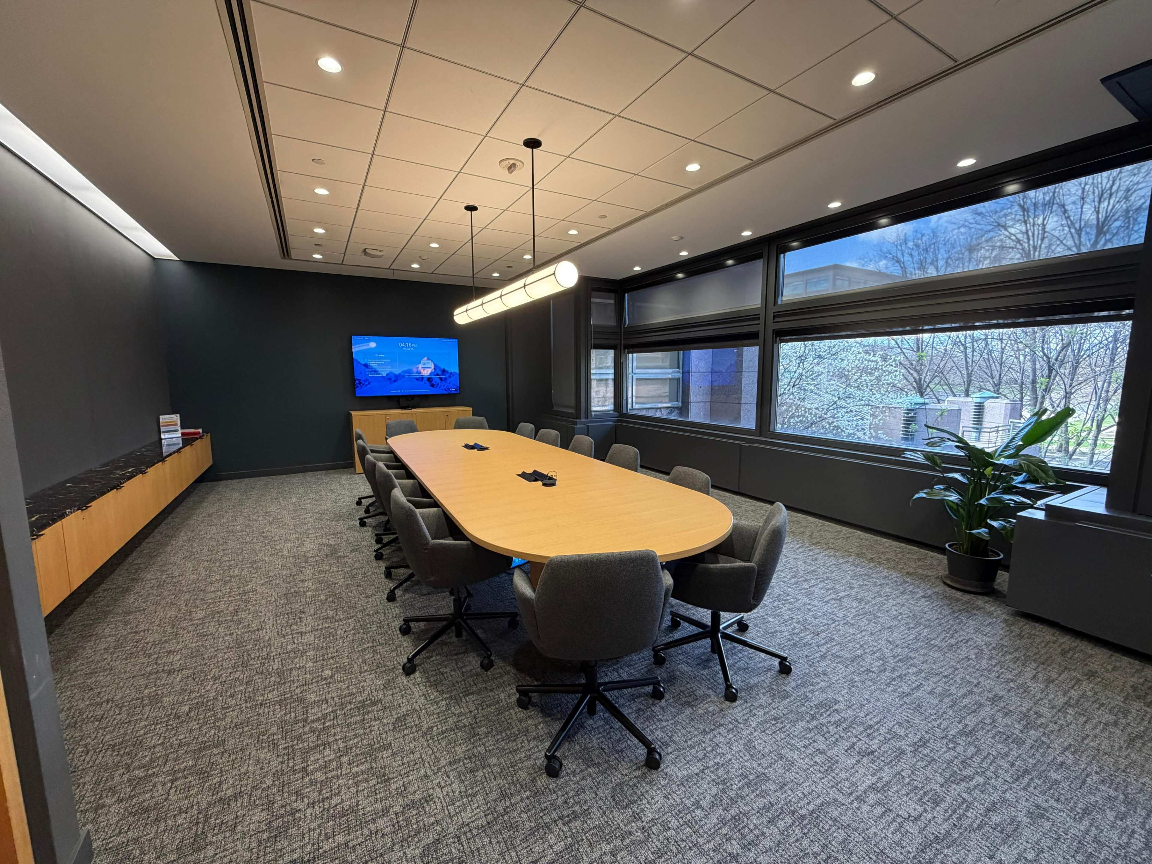 A modern conference room features a long oval table surrounded by rolling chairs, with large windows offering a view of greenery and a screen displaying content.