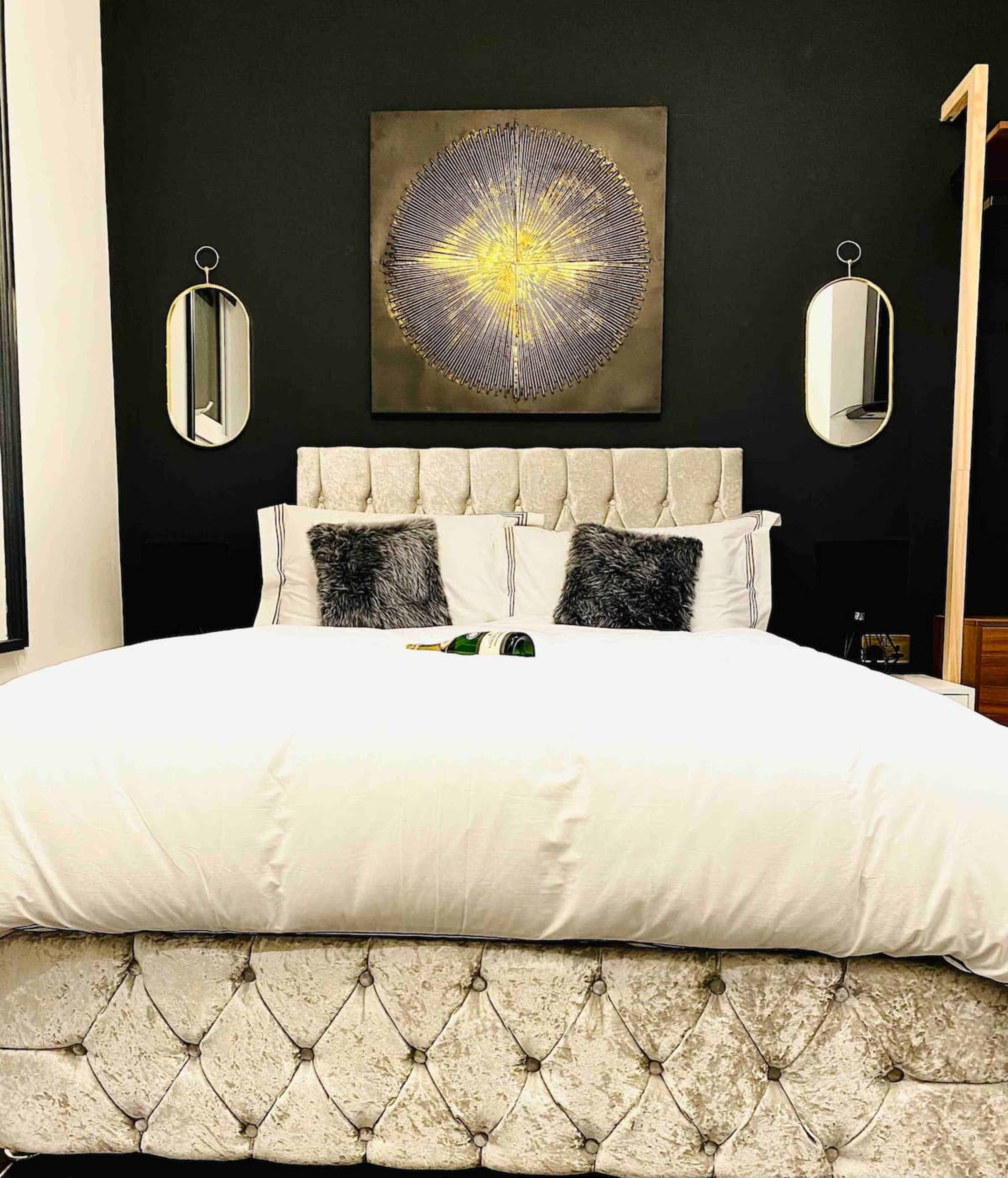 A neatly made bed with a plush upholstered headboard, flanked by two mirrors, against a black wall featuring a geometric artwork.