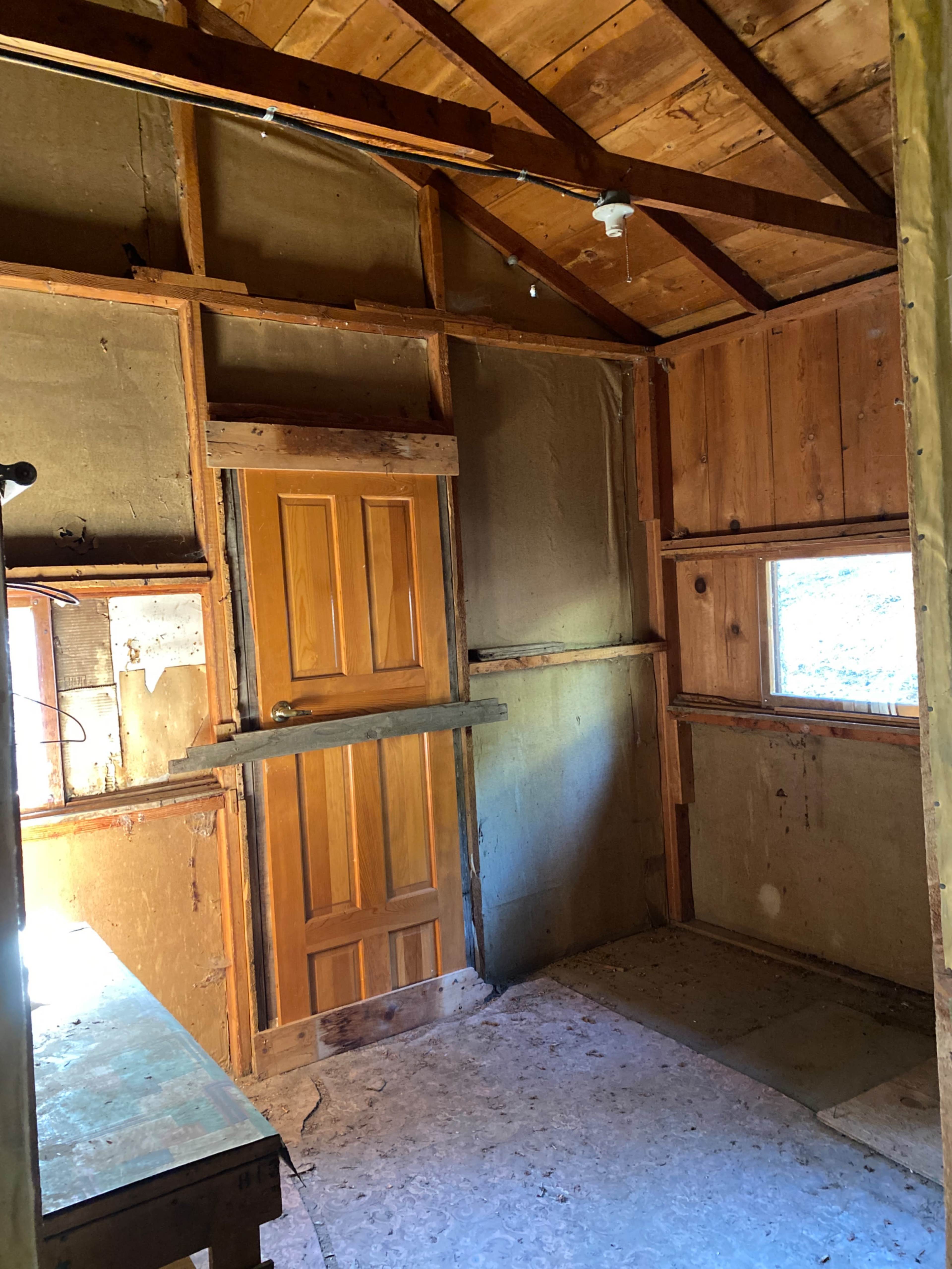 The interior of a rustic, unfinished room features exposed wooden framing, a single door, and a small window.