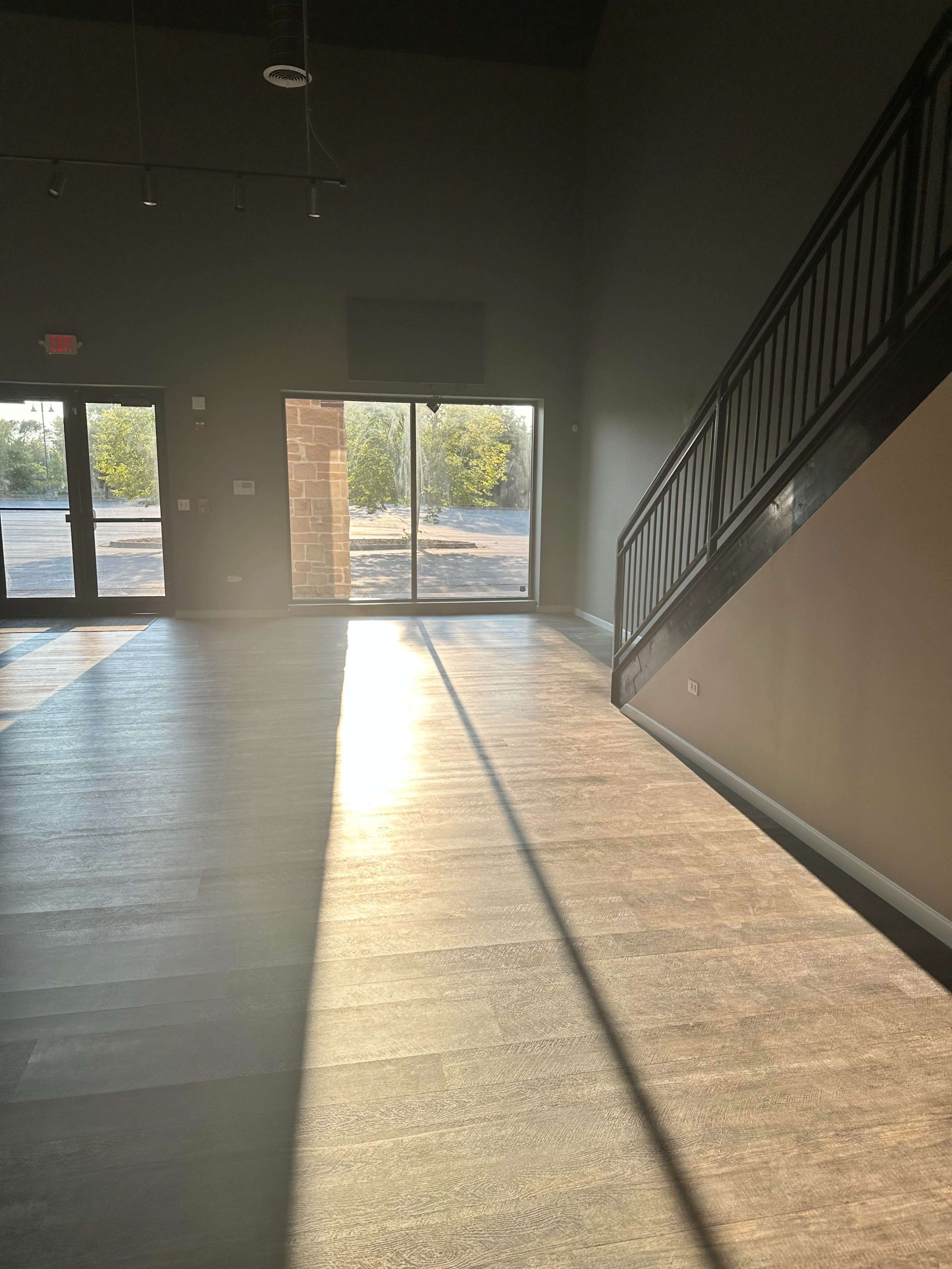 The image shows a spacious interior with a staircase on the right, large windows allowing natural light to enter, and a wooden floor.