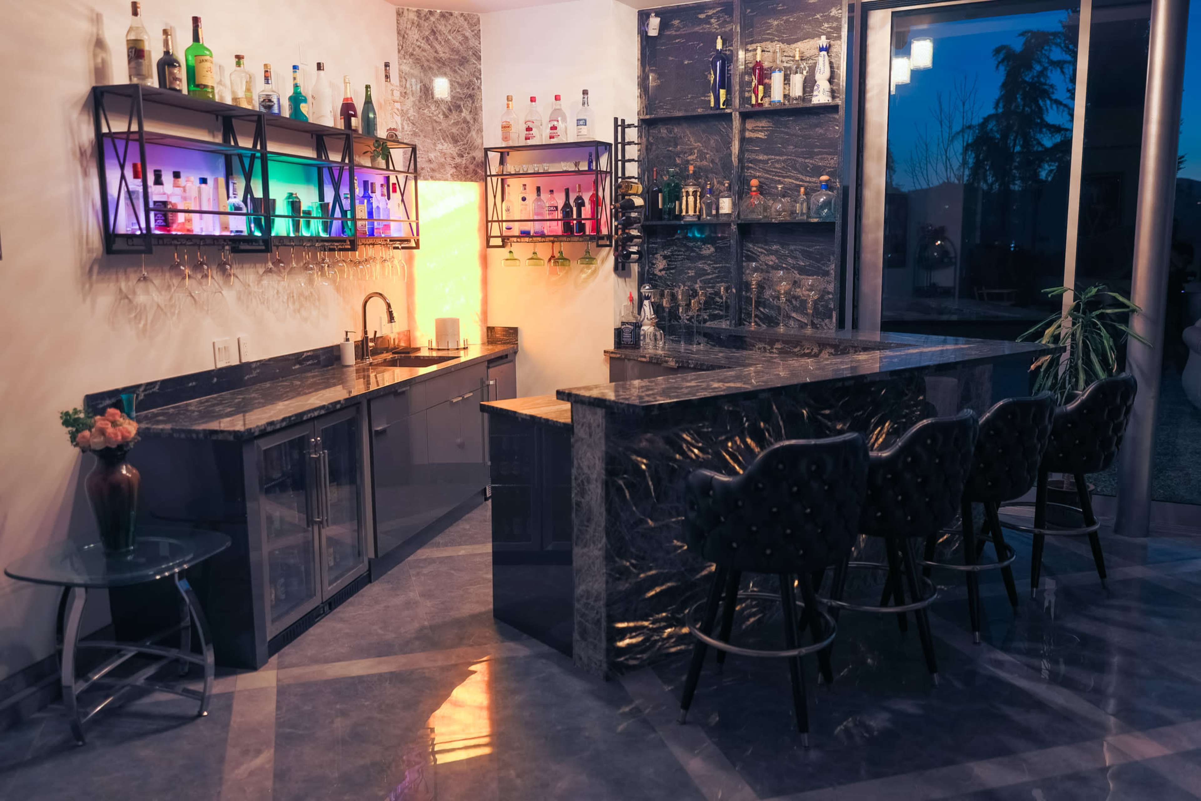 A modern home bar features sleek black marble surfaces, illuminated shelving with various bottles, and stylish seating for four.
