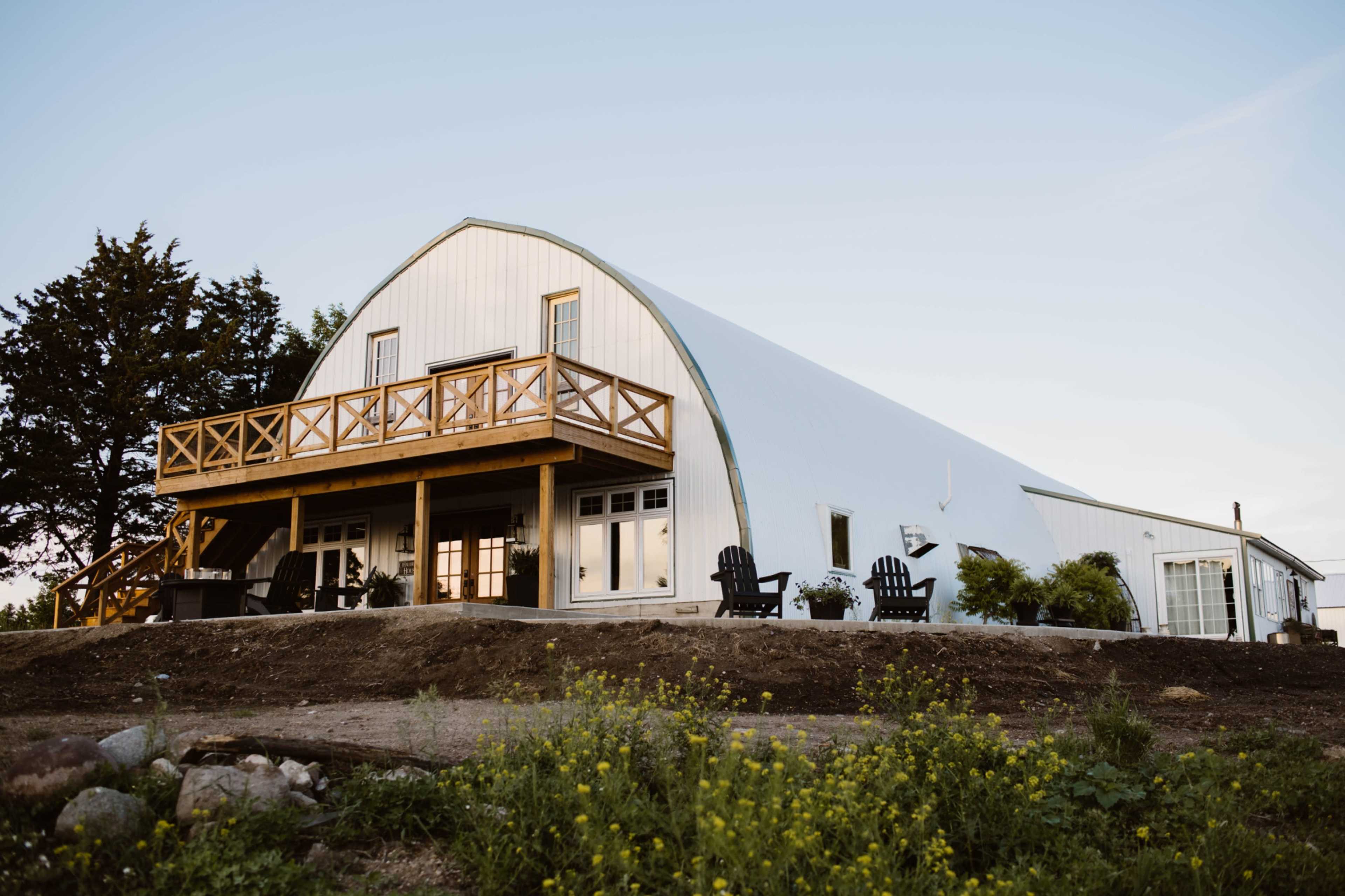 A large, modern barn with a curved roof features a wooden porch and chairs, situated on a grassy hillside.