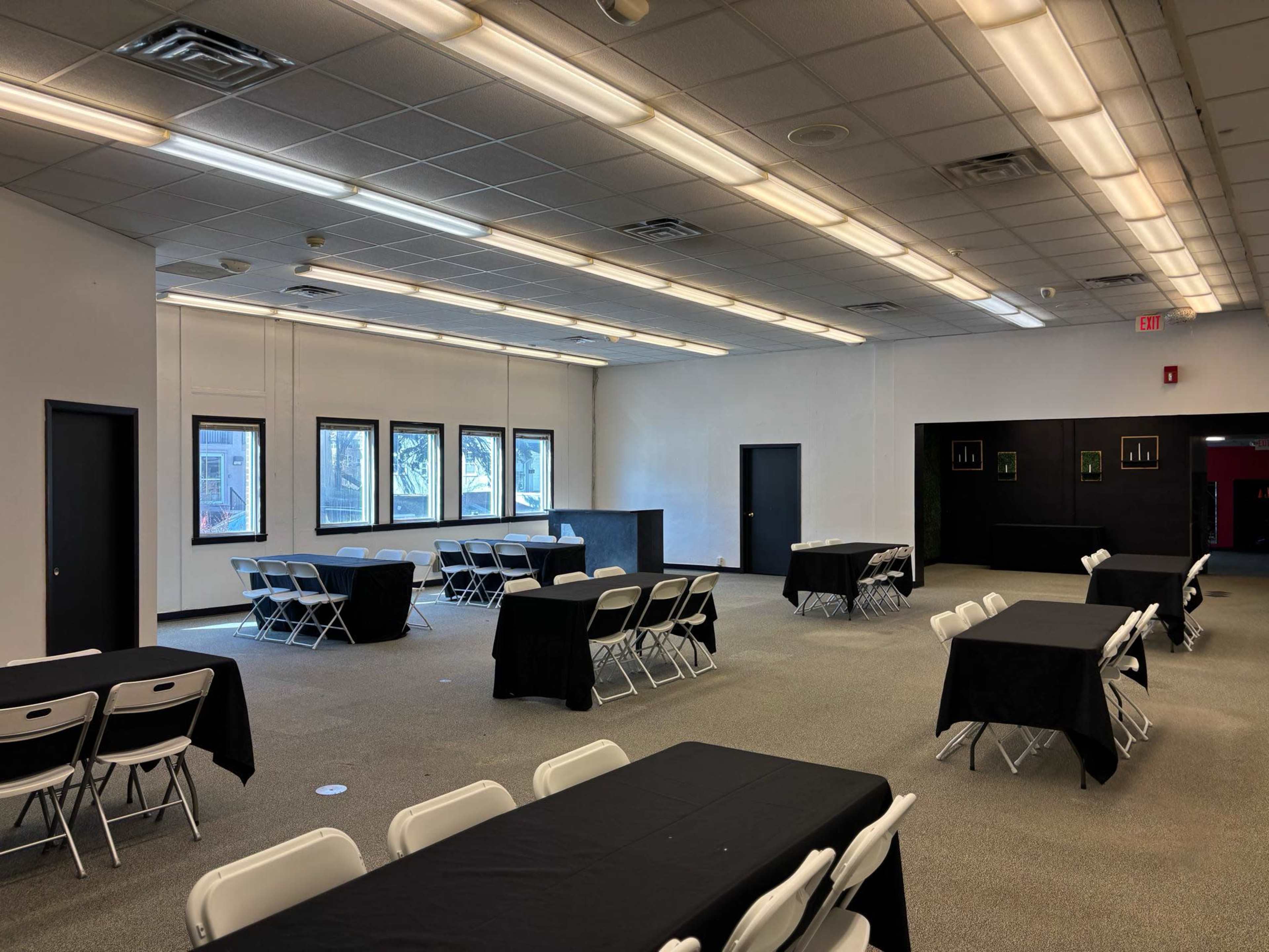The interior of a large, well-lit room features several tables covered with black cloths and arranged with chairs around them, alongside windows and a black counter at one end.