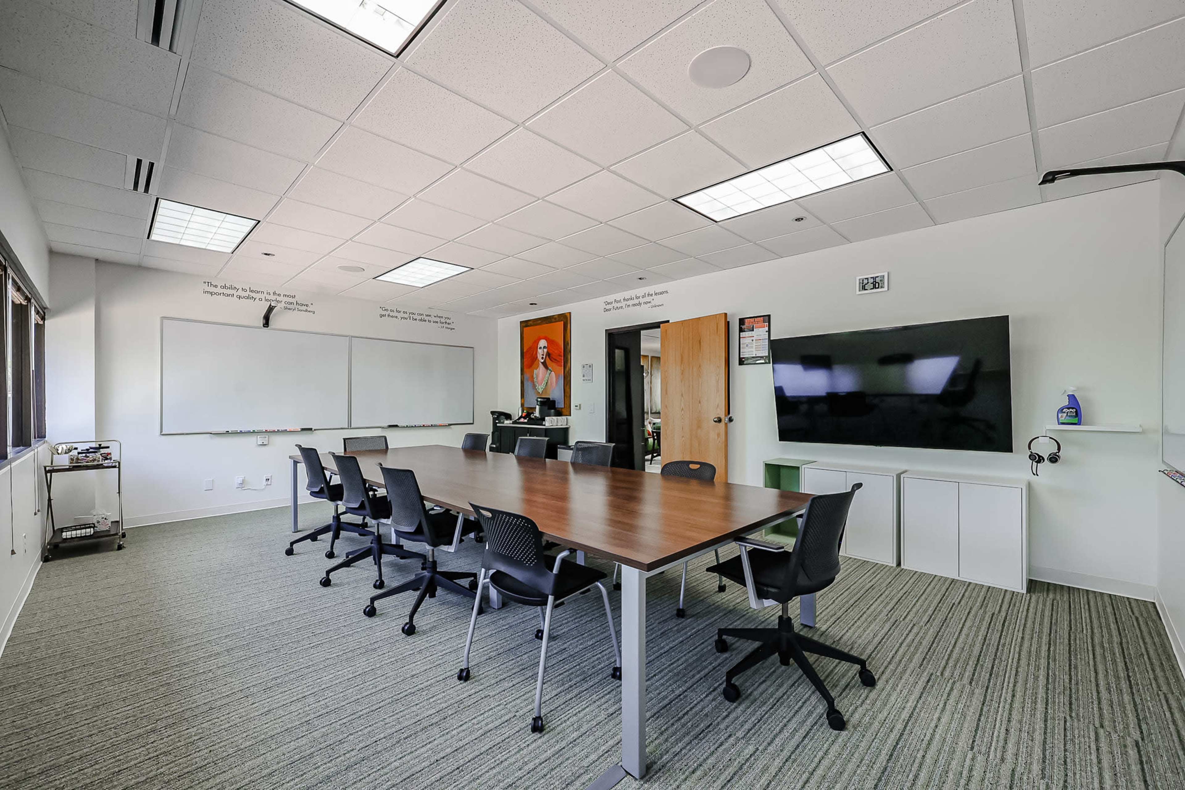 A spacious conference room features a long wooden table surrounded by black chairs, a wall-mounted television, and whiteboards on two walls.
