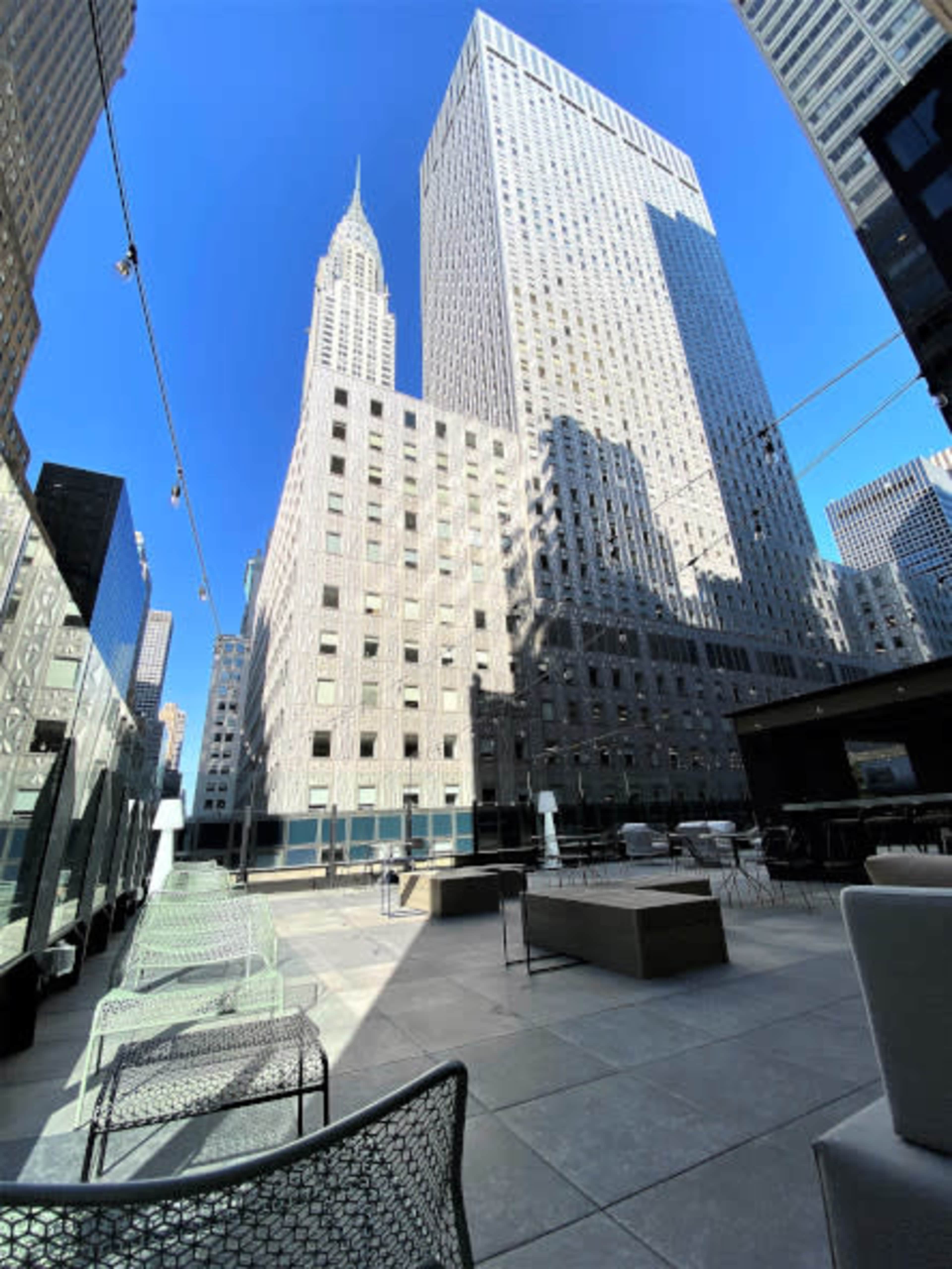 The image shows a modern terrace with seating and a view of tall skyscrapers, including the iconic Empire State Building in the background against a clear blue sky.