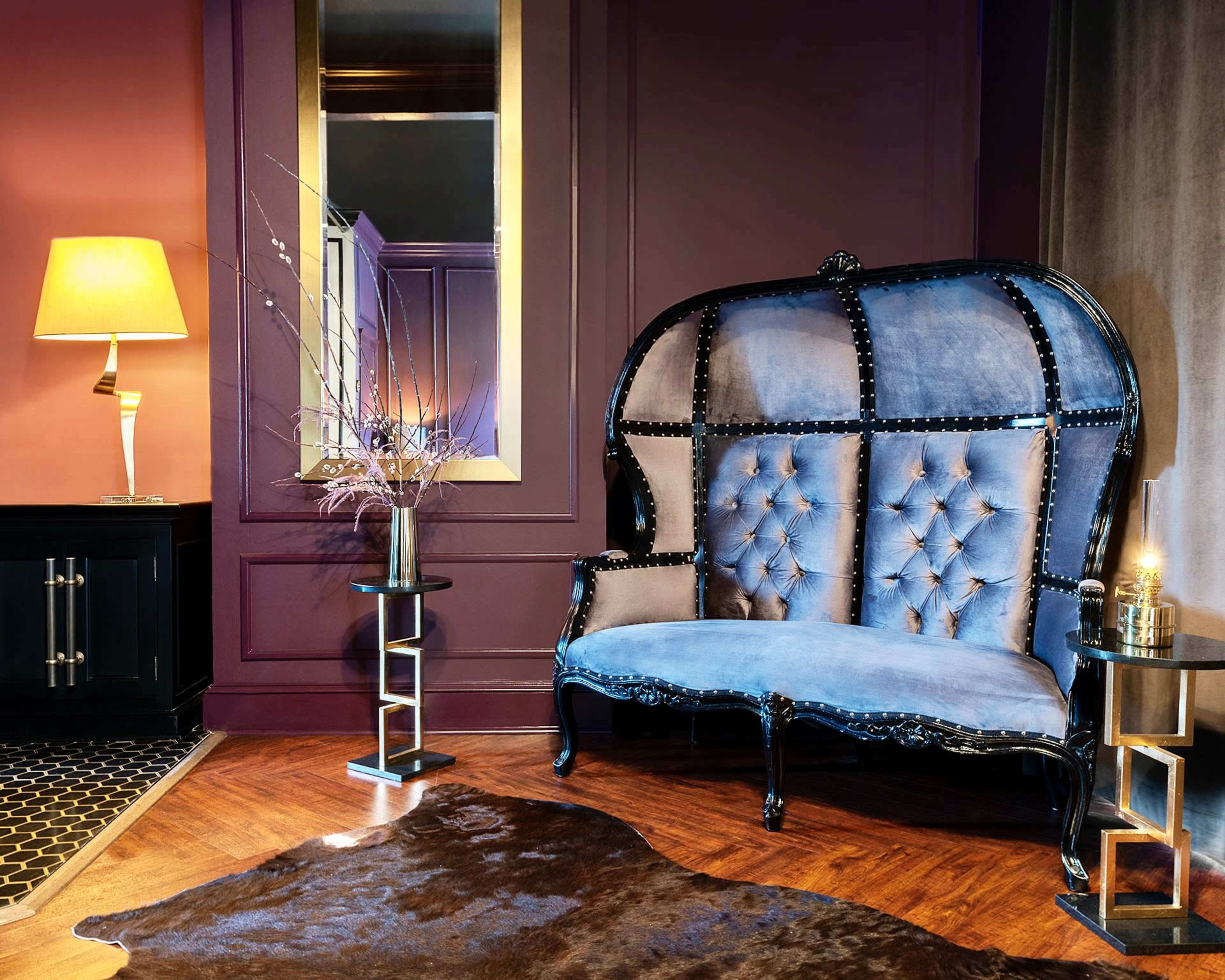 A stylish interior featuring a blue tufted sofa, a lamp, a decorative mirror, and a small side table, all arranged on wooden flooring.