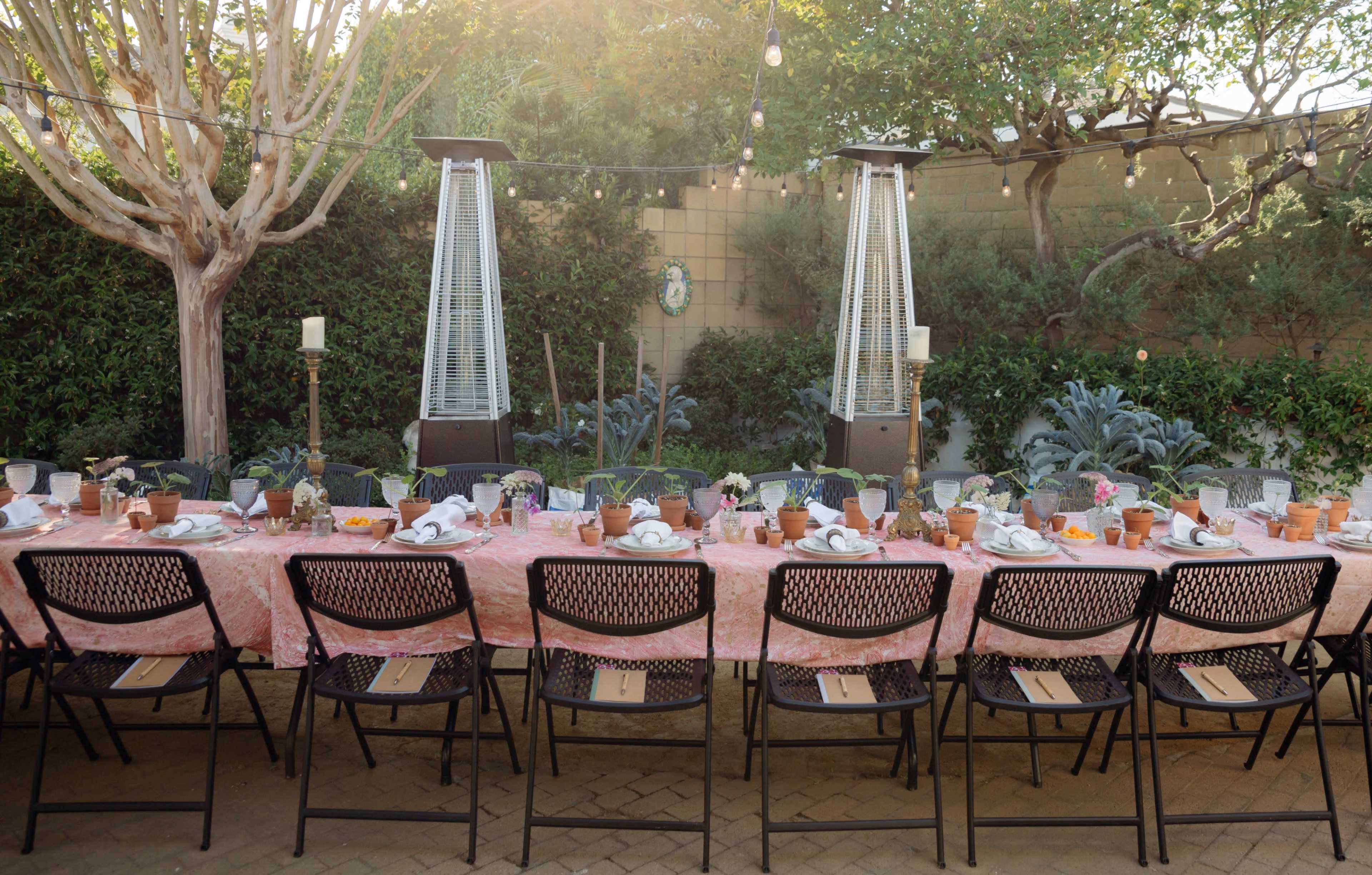 A long outdoor dining table is set with tableware and decorated with potted plants, surrounded by greenery and patio heaters.