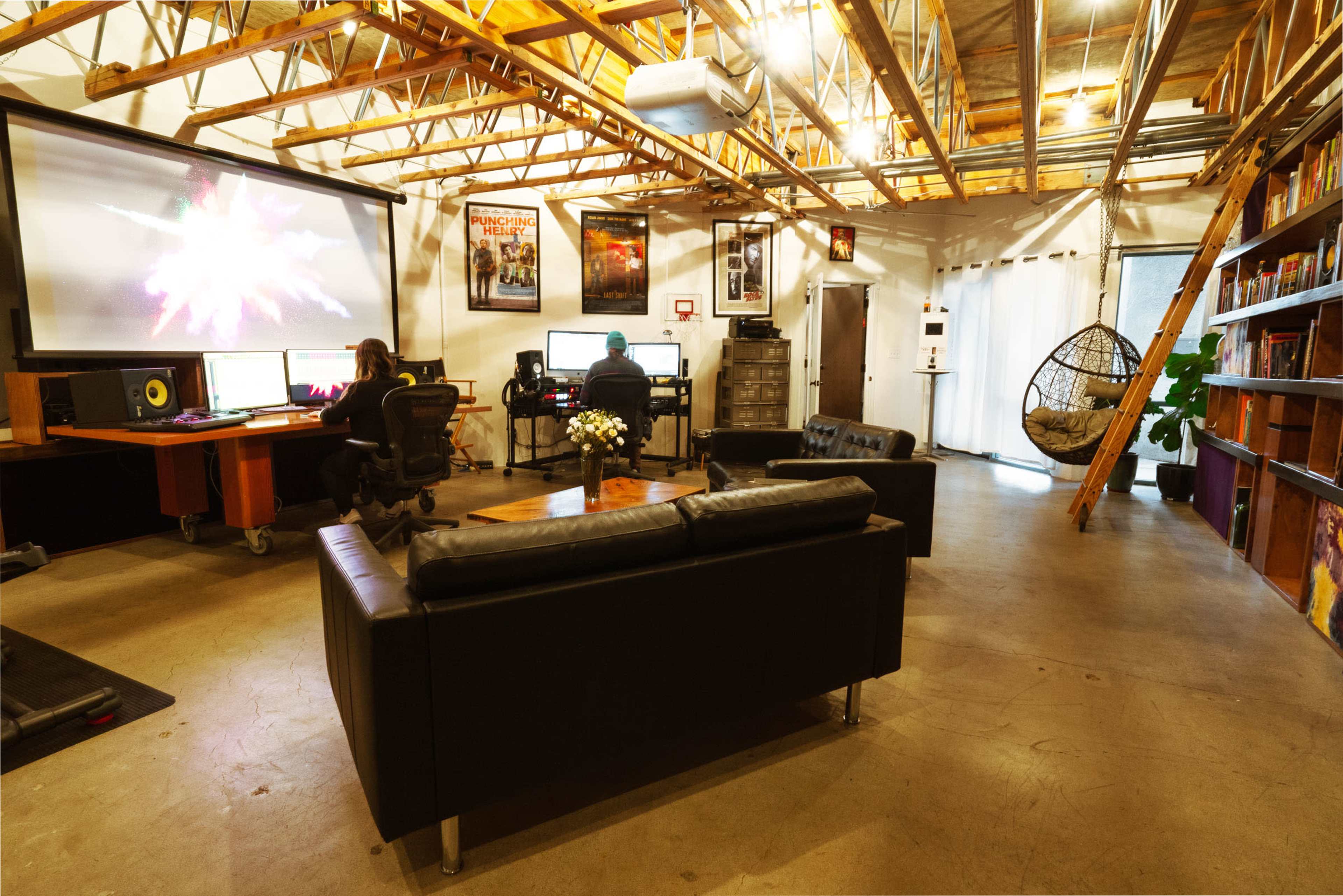 A spacious, modern office with two desks, a large screen displaying visuals, a black leather sofa, and a hanging chair in a well-lit area.