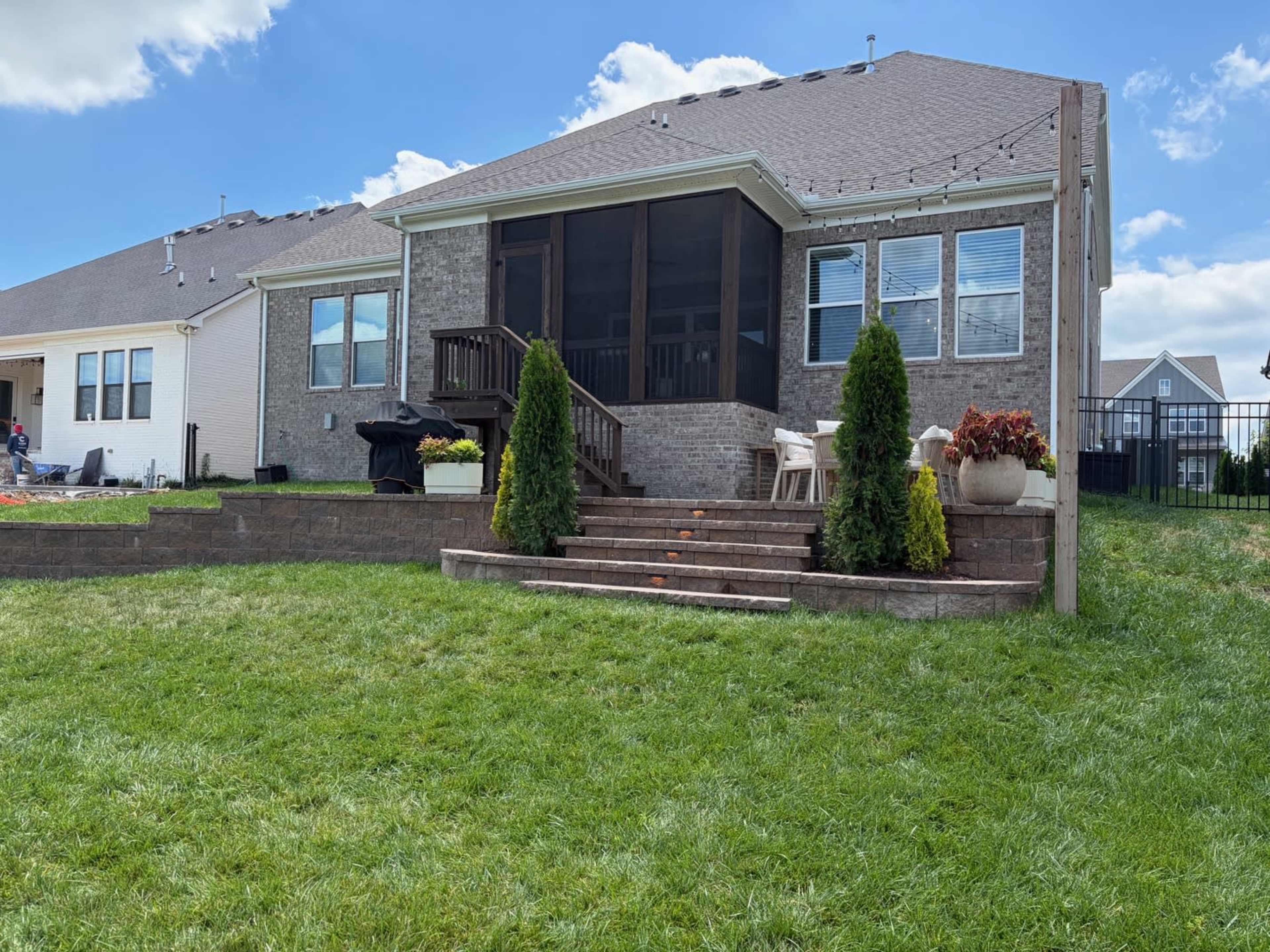 A backyard features a two-story brick house with a screened porch, steps leading to a grassy area, and decorative plants on a sunny day.