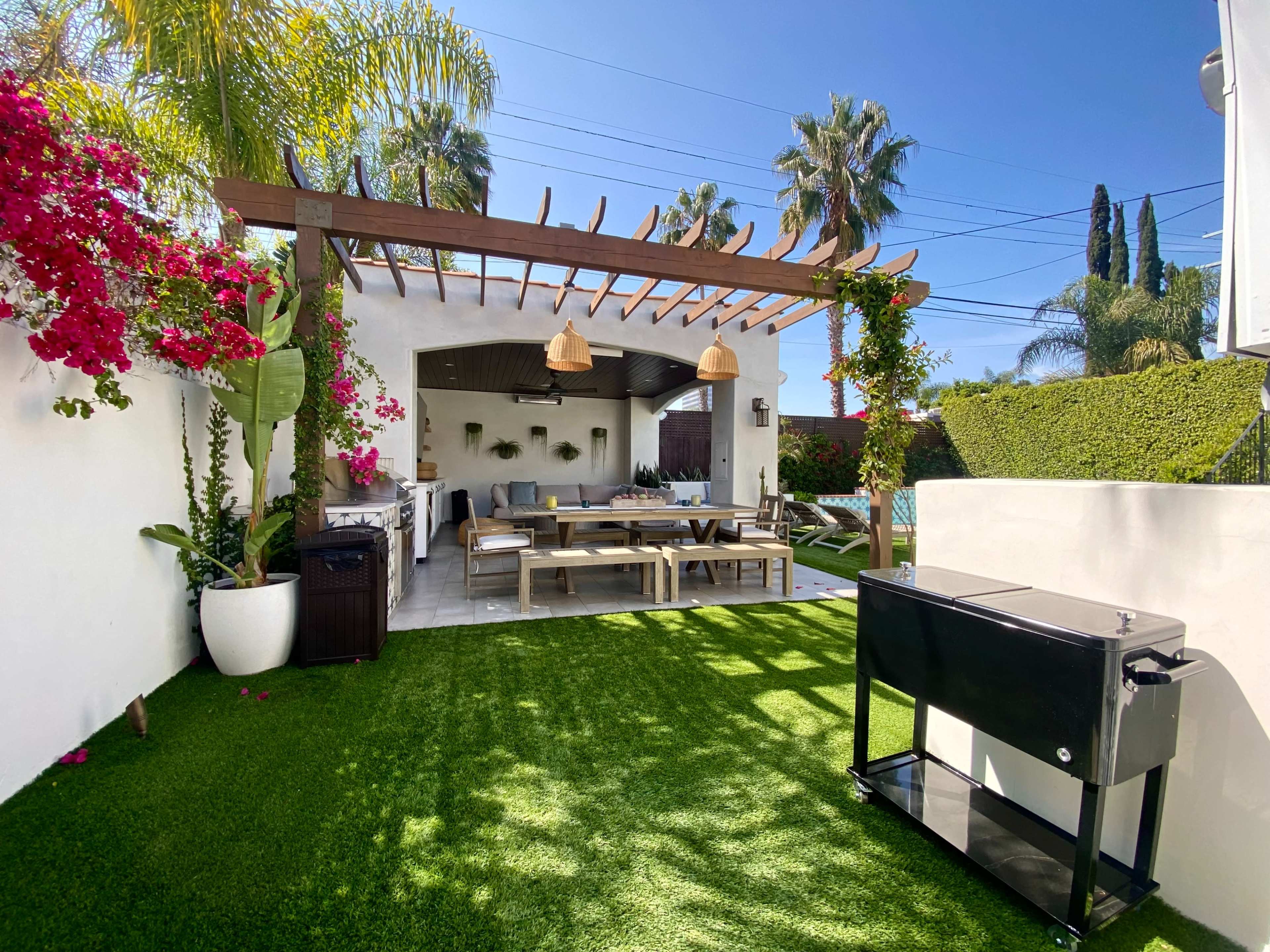 The image shows a landscaped backyard with a seating area under a pergola, surrounded by greenery and flowering plants.