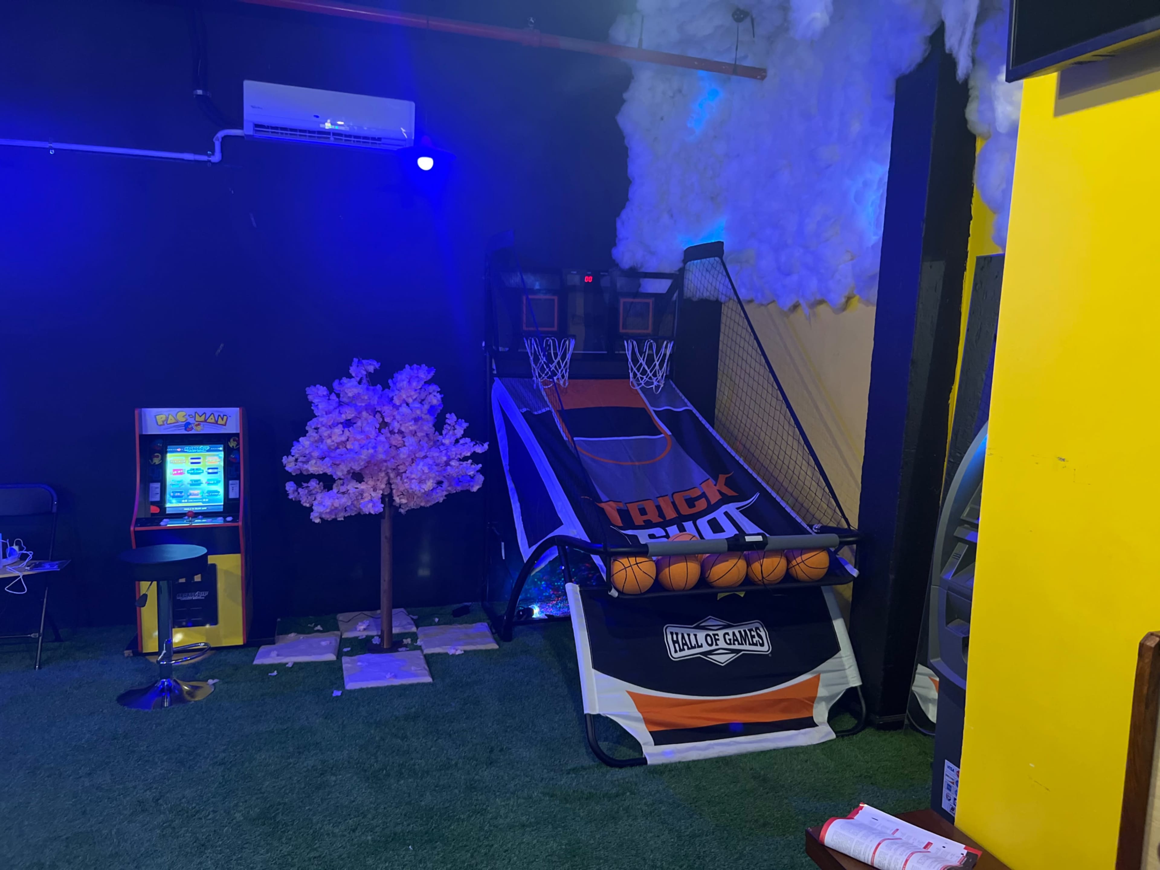 An indoor arcade featuring a basketball shooting game, a small tree decoration, and a vending machine against a backdrop of dark walls and colorful lighting.