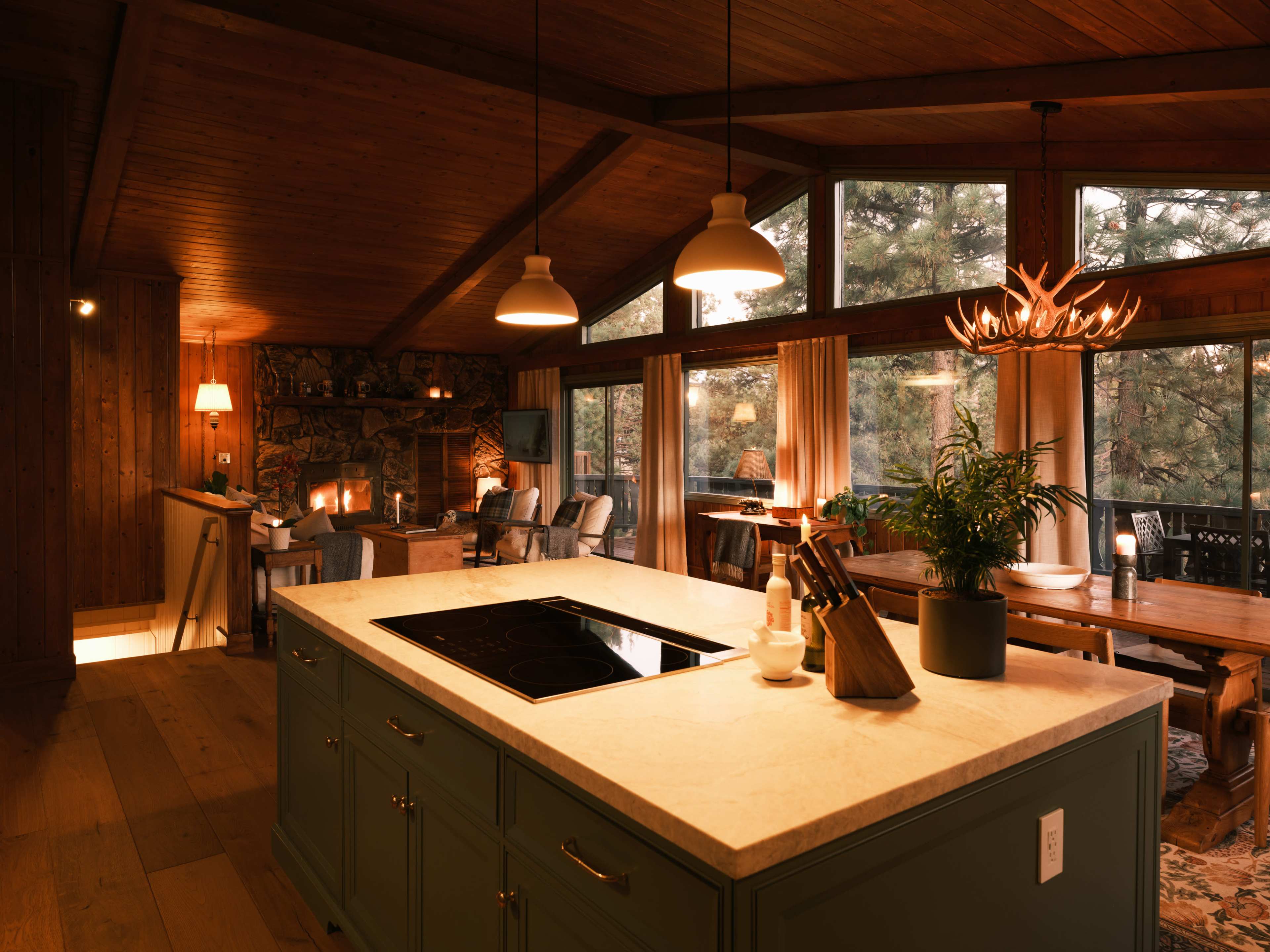 A modern kitchen with an island features a cooktop, surrounded by a dining area and living space with large windows overlooking a forest.