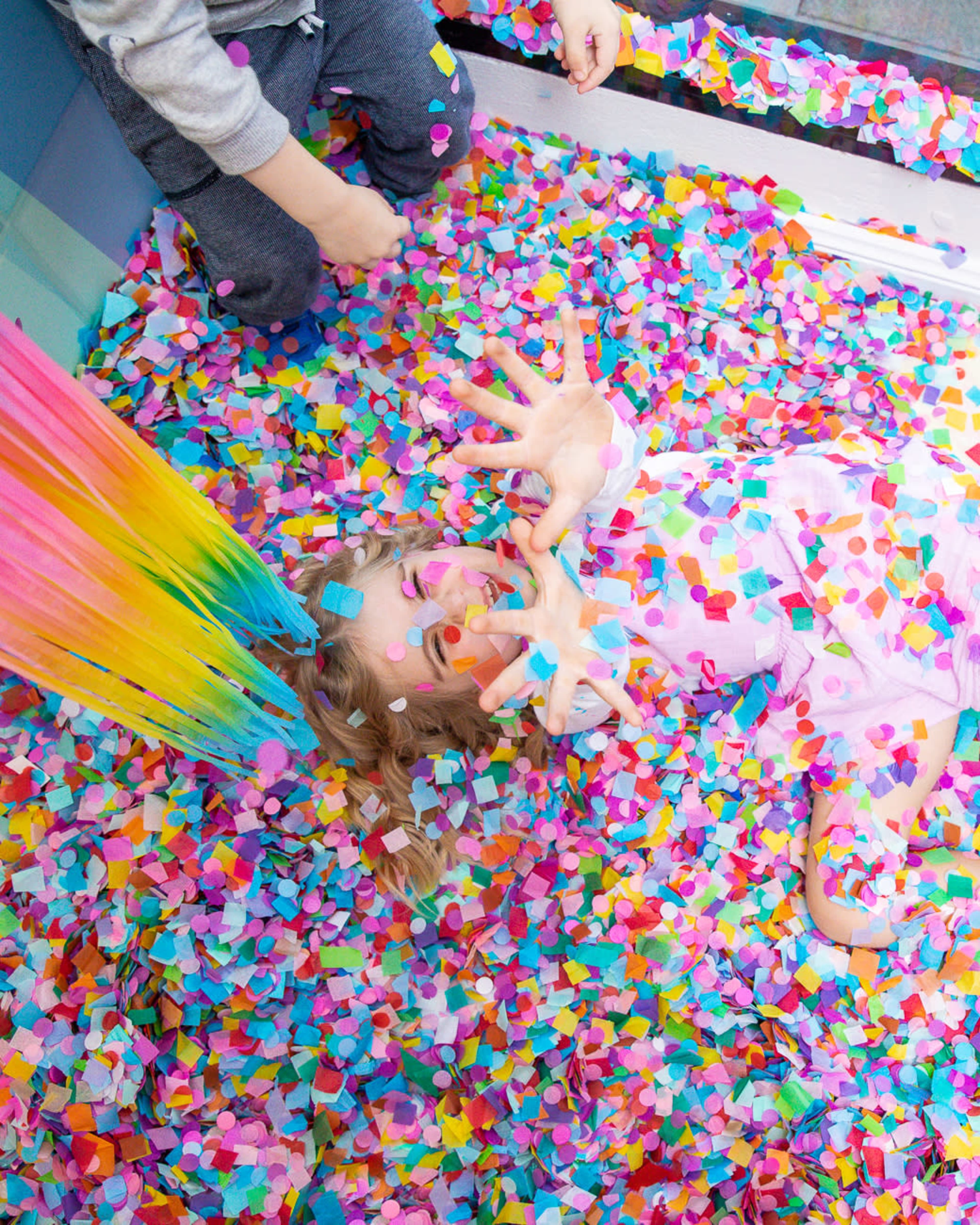 A child lies among a bed of colorful confetti, surrounded by vibrant strands of tissue paper.