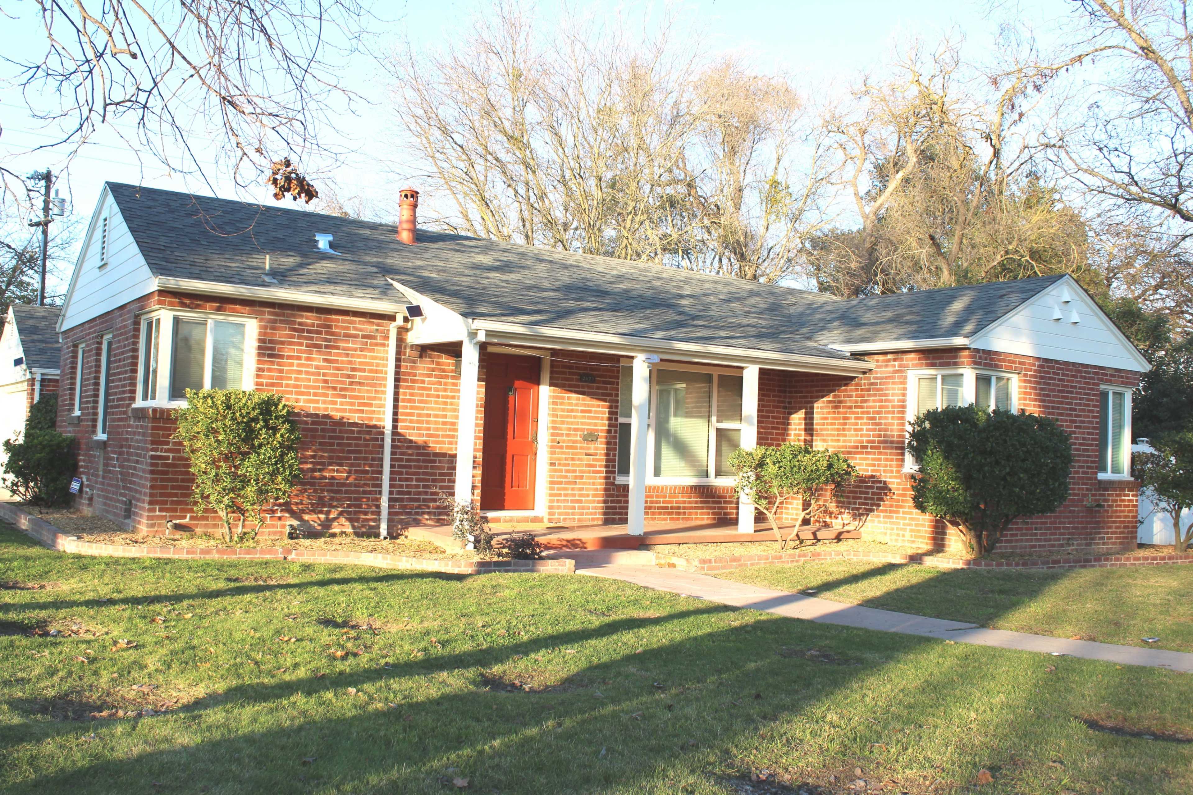 A brick bungalow with a gray roof has a front porch and neat landscaping, situated on a grassy lawn.