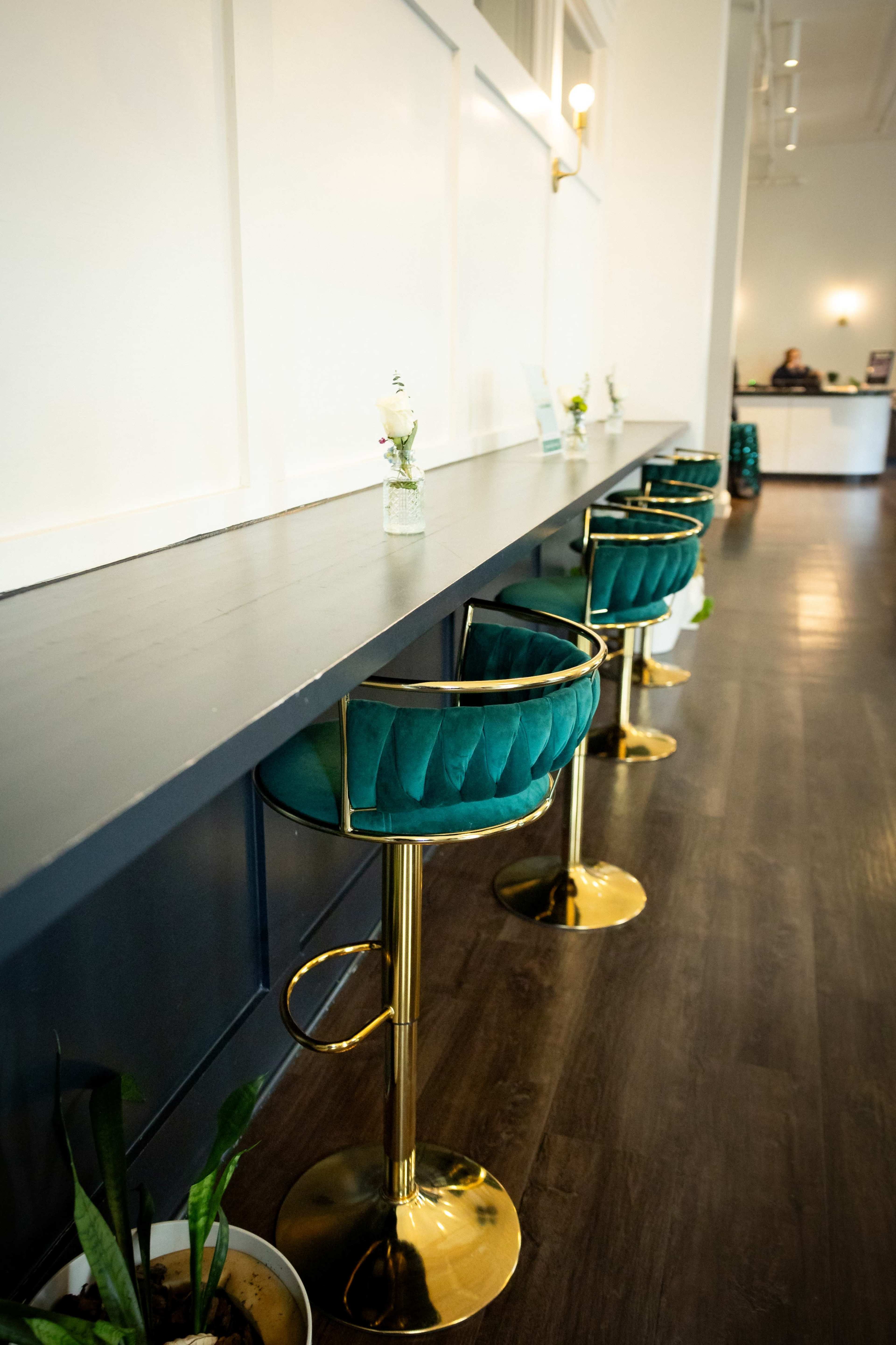 A row of gold and teal bar stools is positioned along a long dark countertop, with small vases placed on the surface.
