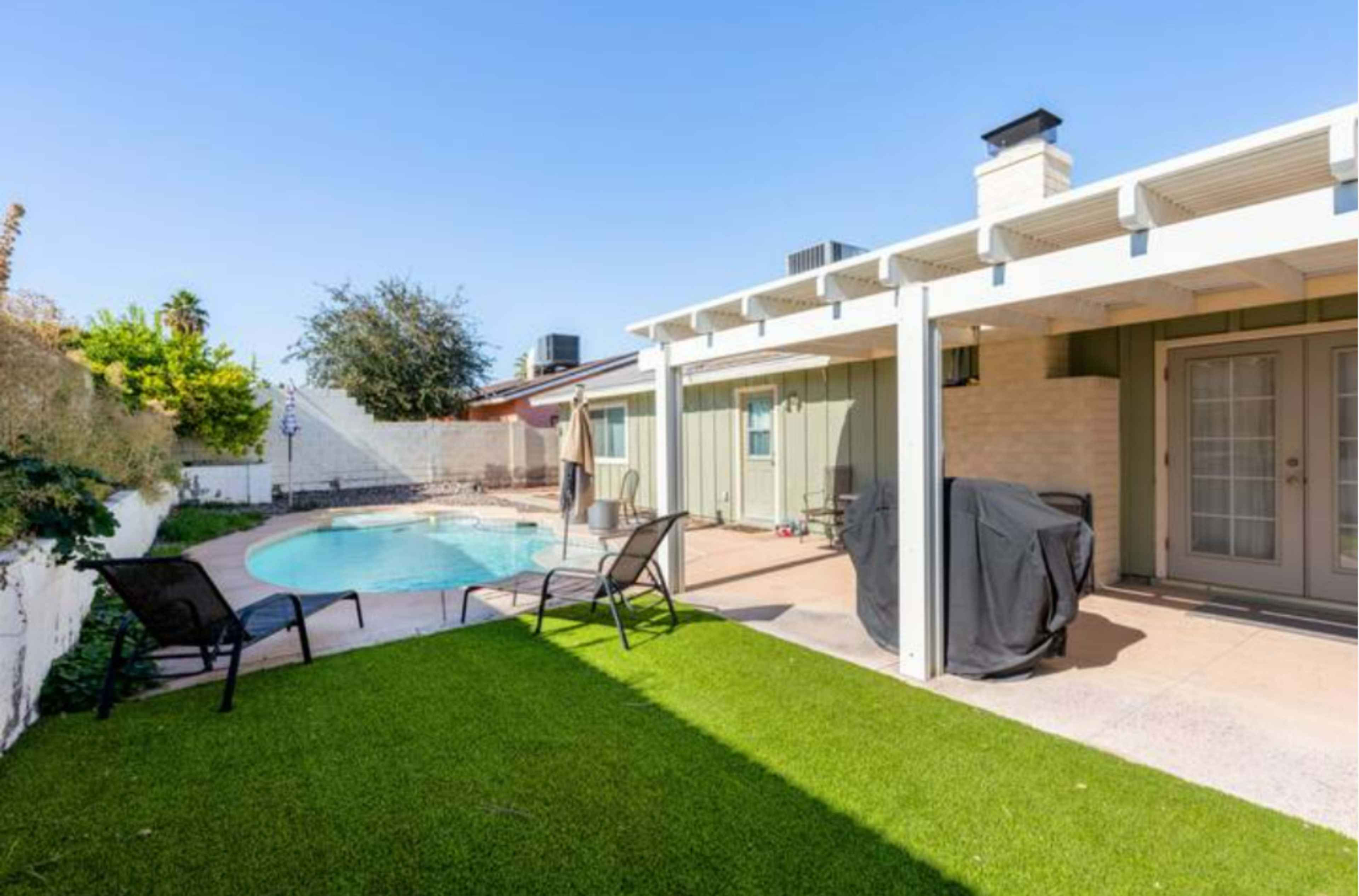 The image shows a backyard with a swimming pool, a covered patio, and outdoor seating on green grass.