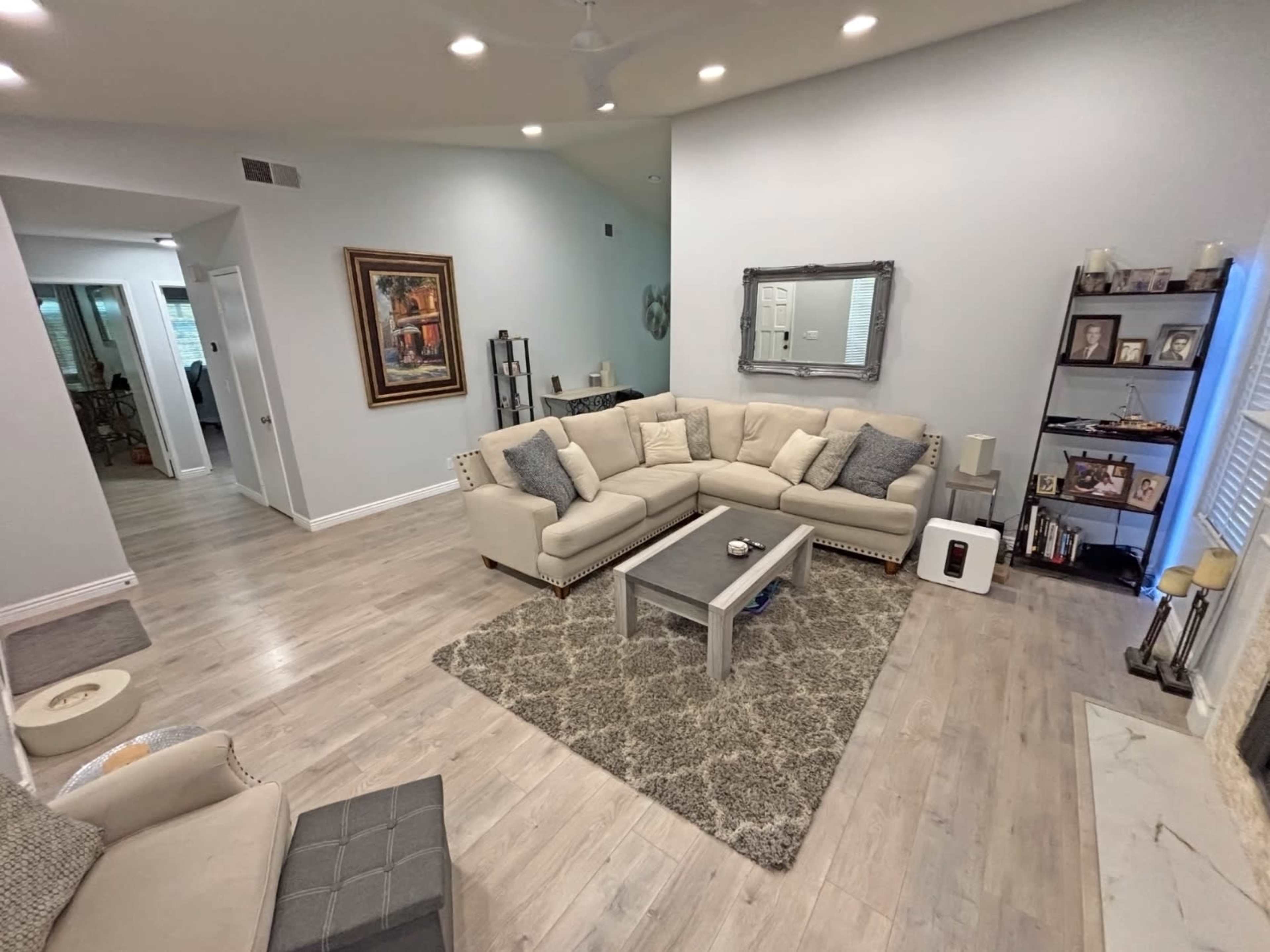 The image shows a spacious living room with light-colored walls, a beige sectional sofa, a coffee table, and a decorative rug on a wooden floor.