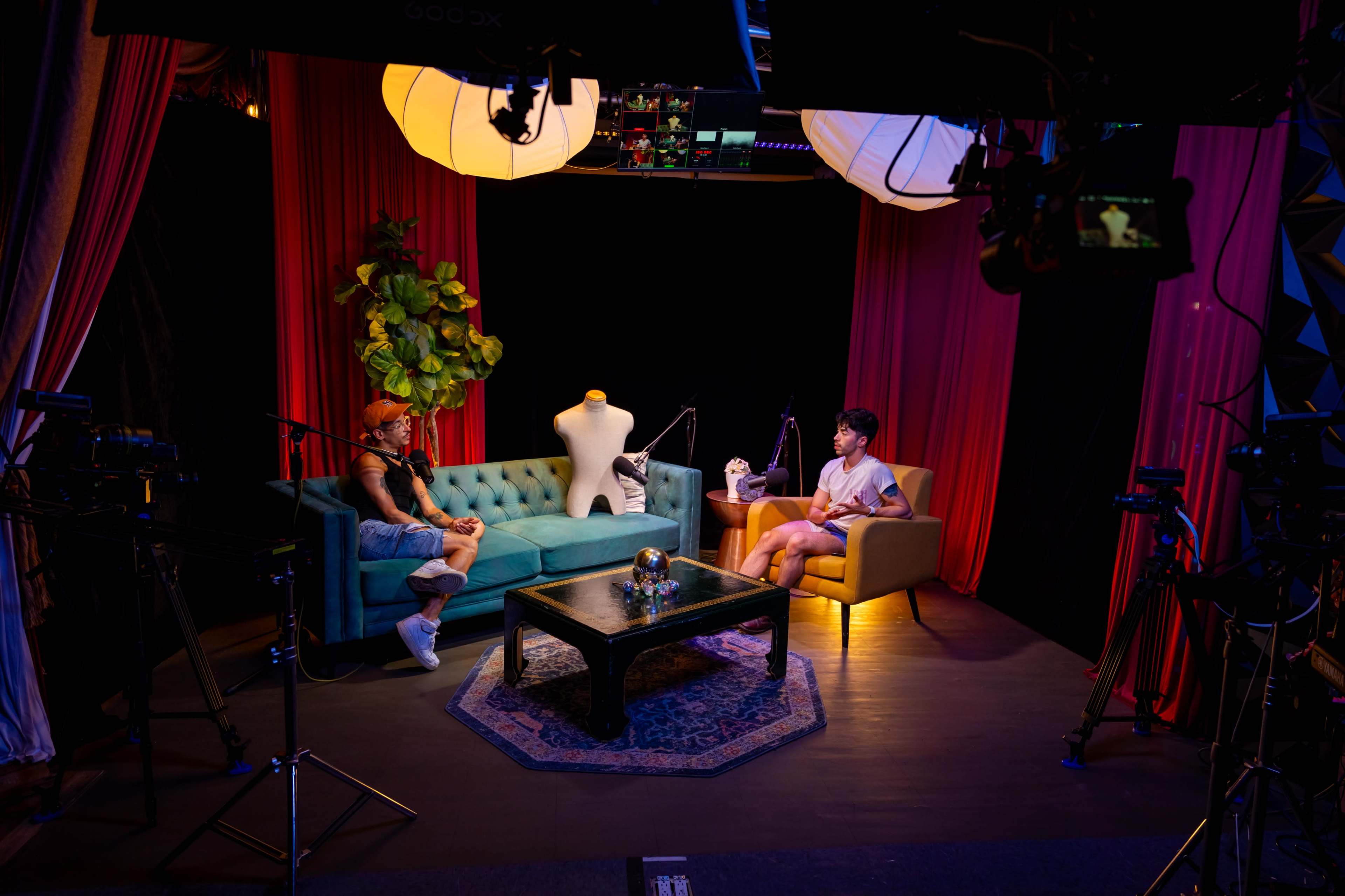 A talk show set features two people seated on a teal sofa and a beige chair, with cameras and lighting equipment positioned around them.