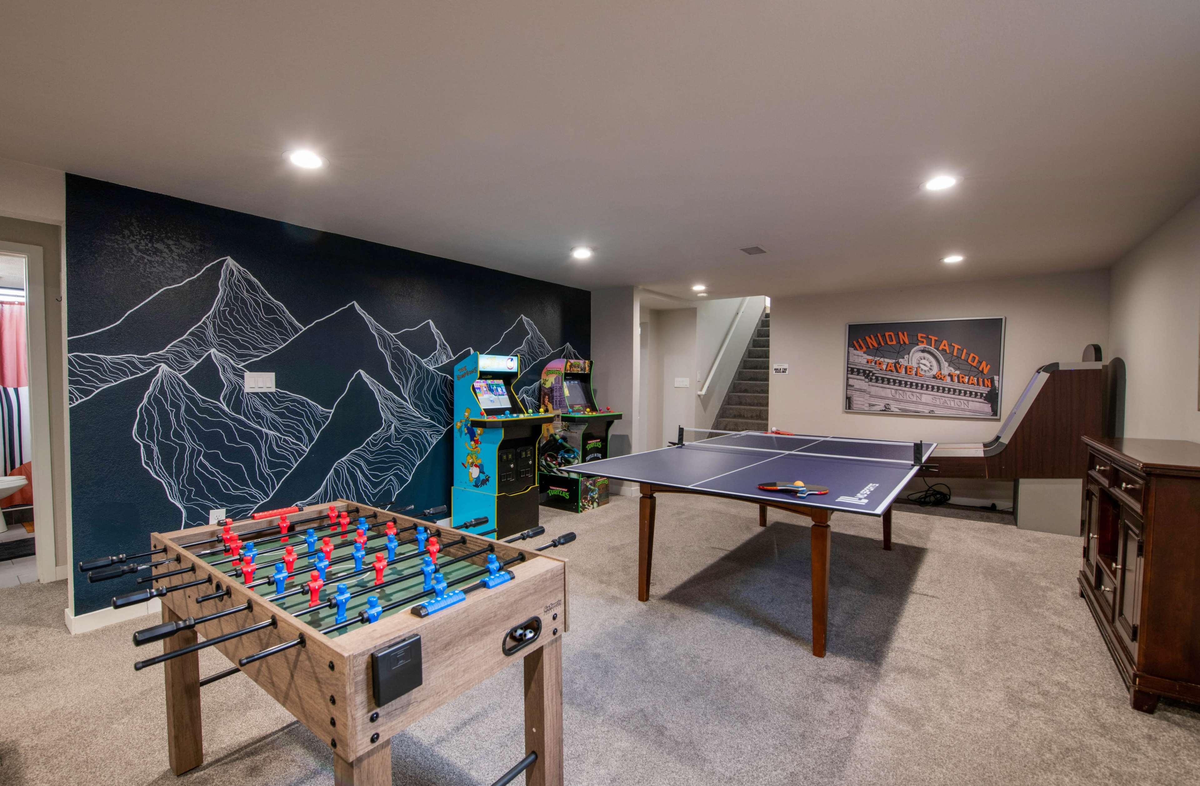 The image shows a basement game room featuring a foosball table, an arcade machine, a ping pong table, and a mountain mural on the wall.