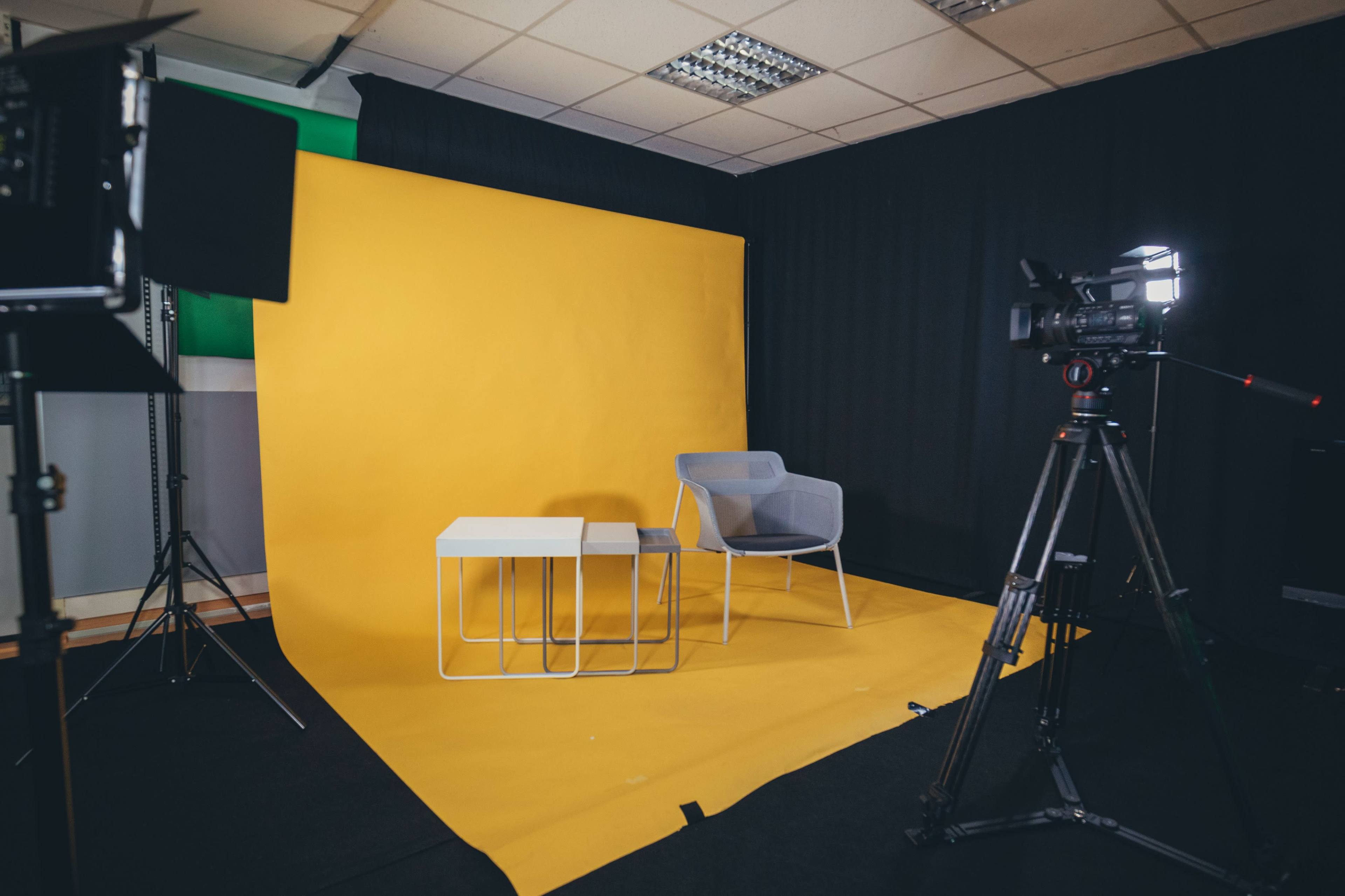 A photography or video studio setup with a yellow backdrop, a gray chair, two white tables, and equipment for recording on the right.