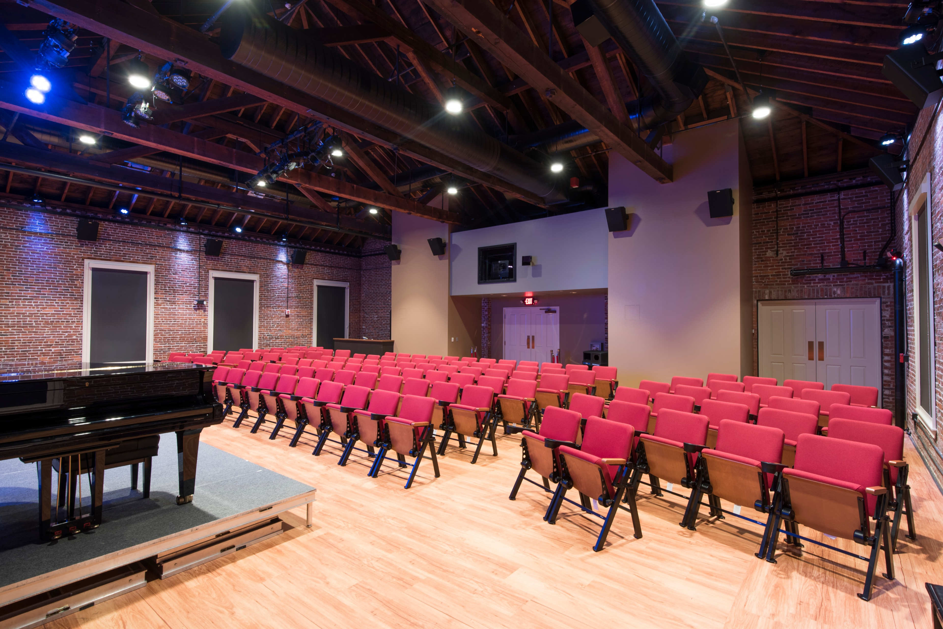 A concert hall with red seats arranged in rows facing a black grand piano on a stage, illuminated by overhead lights.