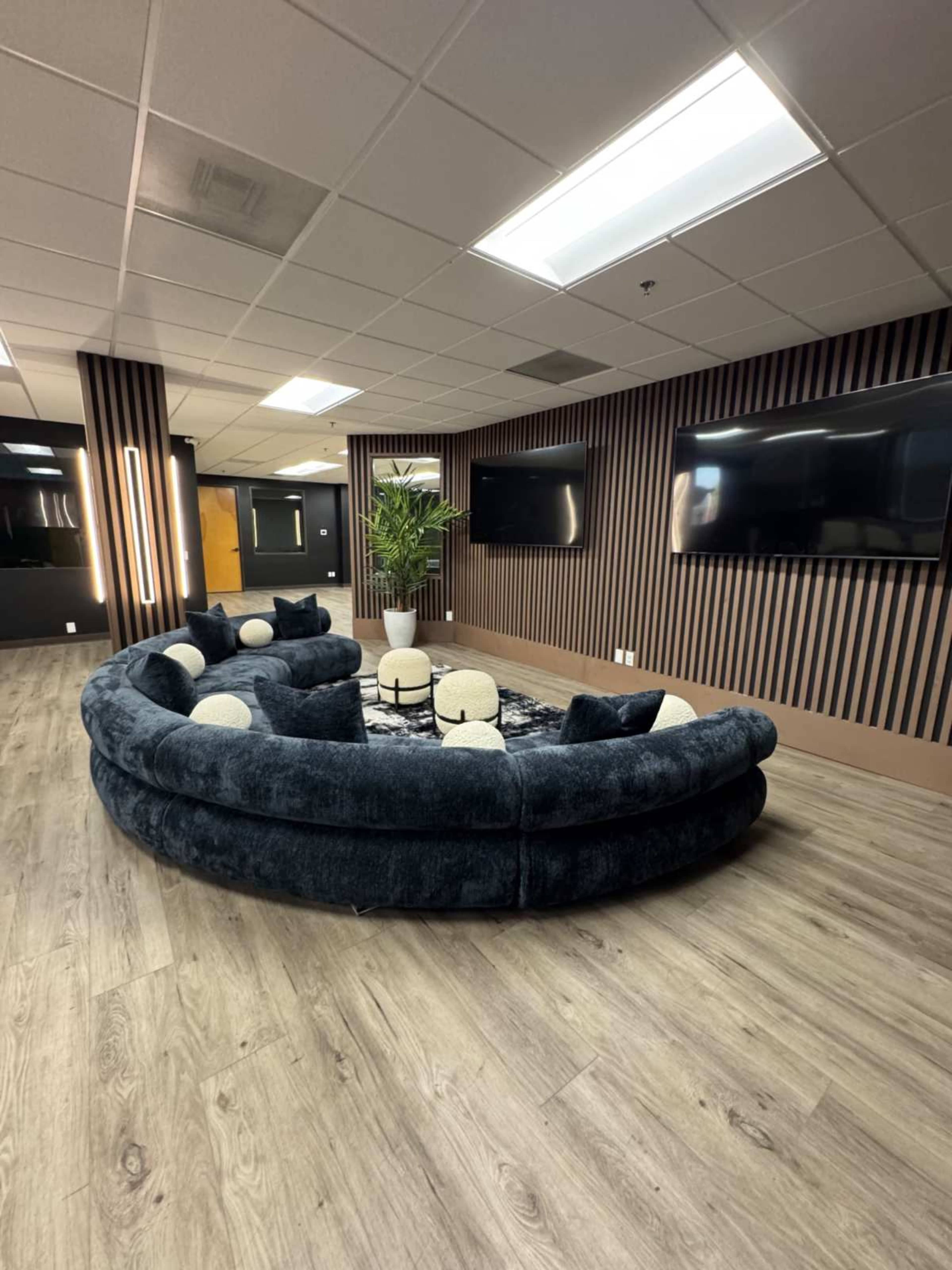 The image shows a modern lounge area featuring a large circular couch with decorative pillows, surrounded by wall-mounted television screens and wooden flooring.