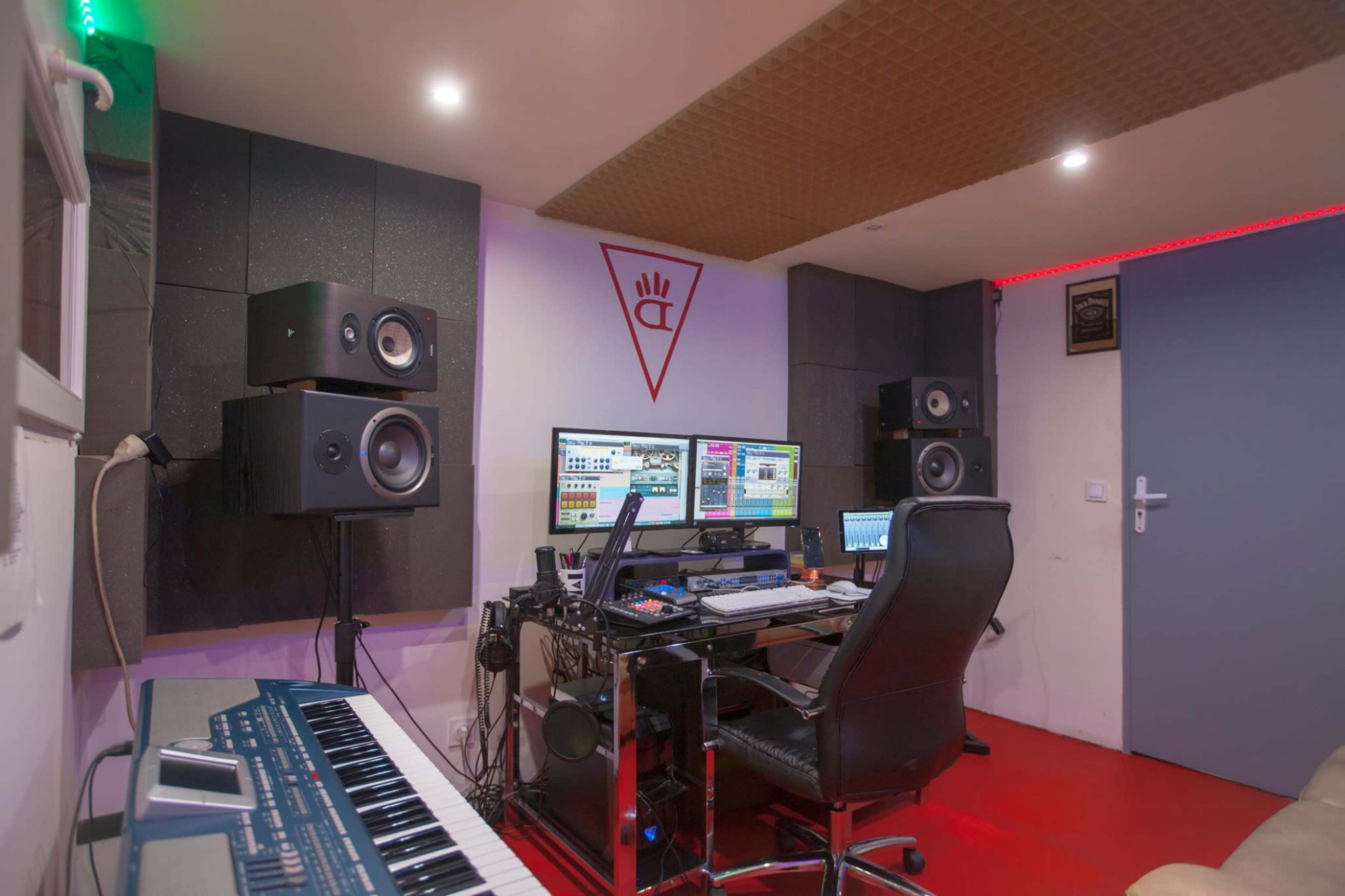A music production studio featuring a desk with two computer monitors, studio speakers, a keyboard, and soundproofing on the walls.