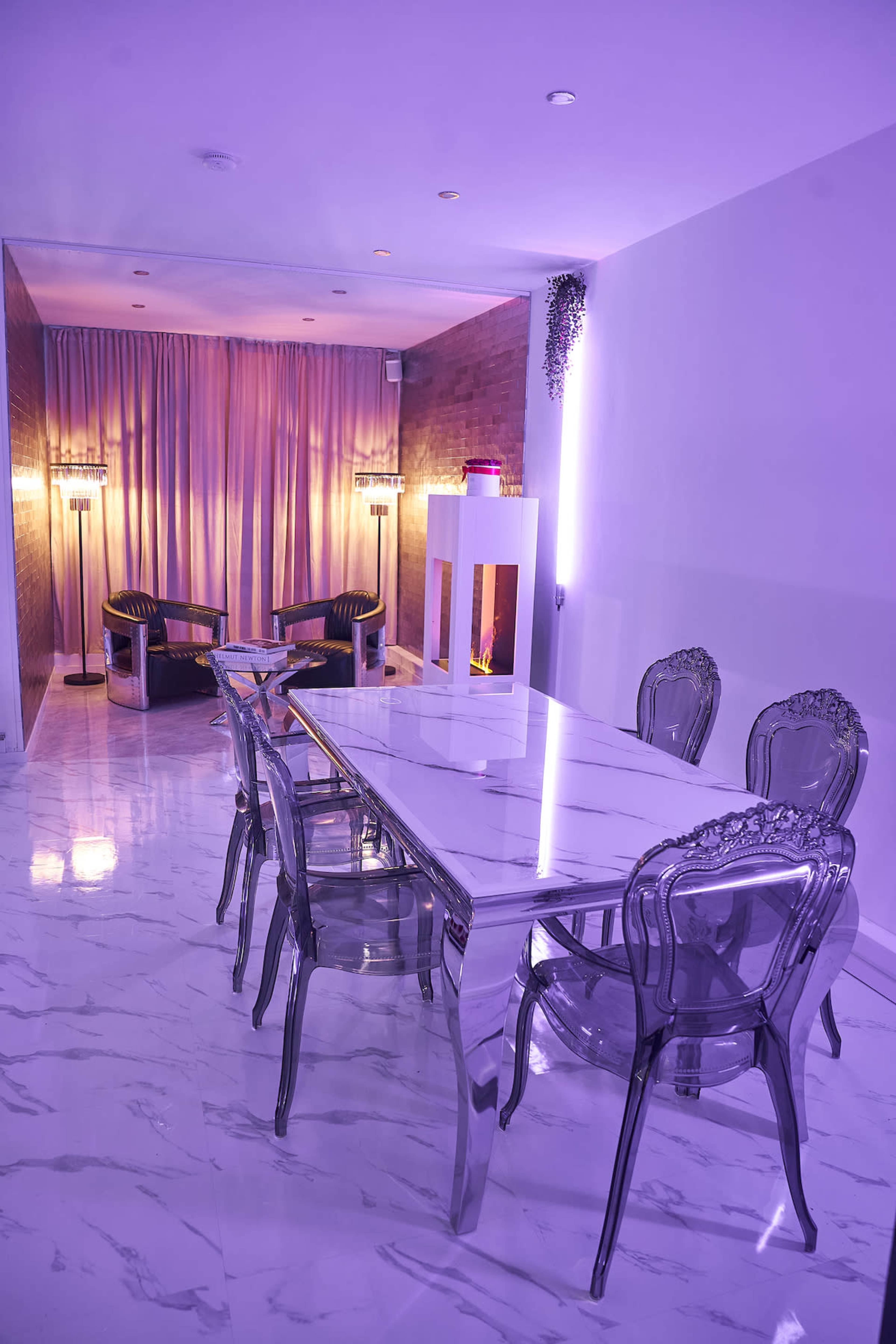 A modern dining area features a glossy white table with clear chairs, accentuated by purple lighting and a cozy seating area in the background.