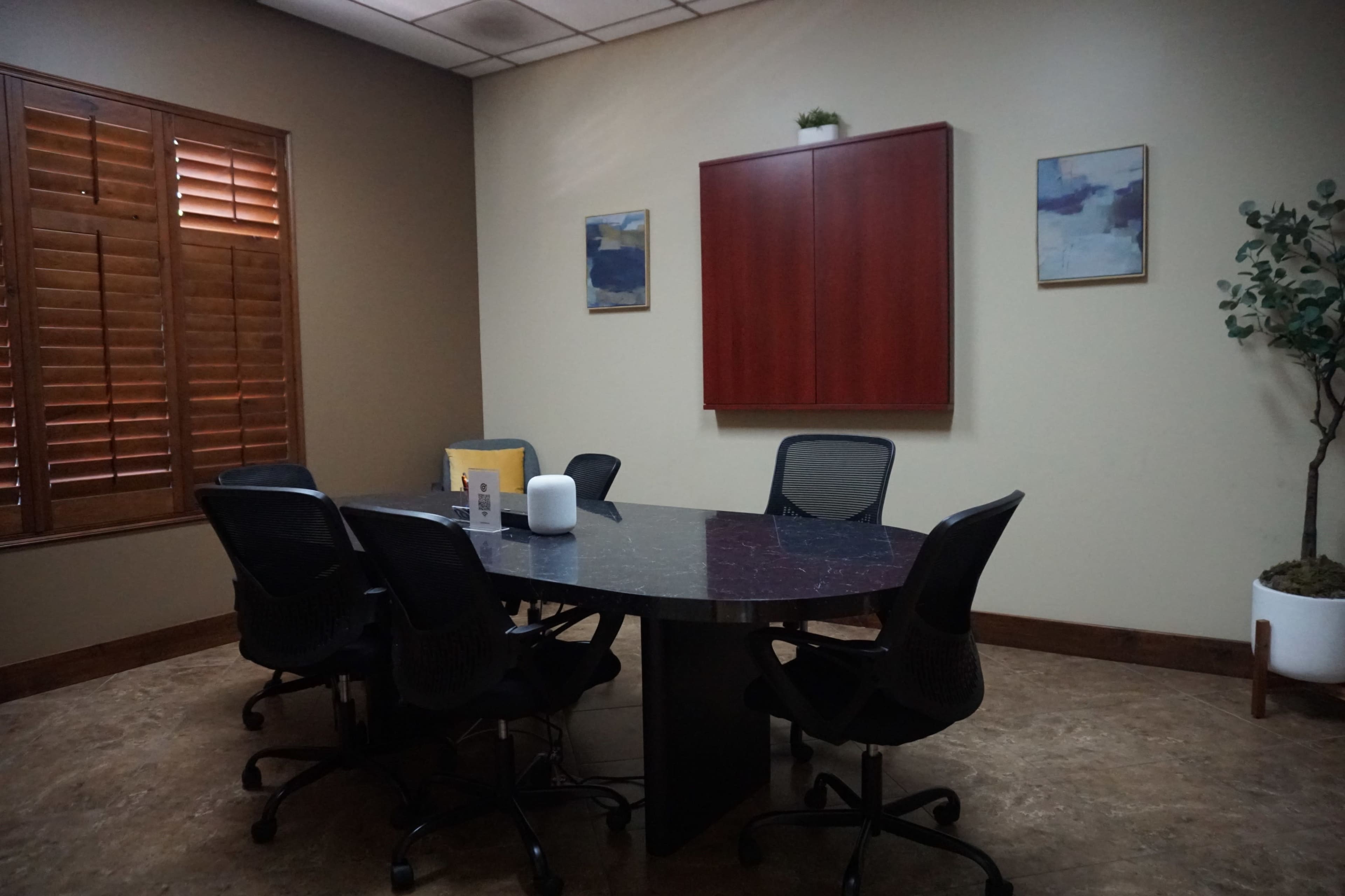 A conference room features a round table surrounded by six black ergonomic chairs, with wooden blinds and two framed artworks on the walls.
