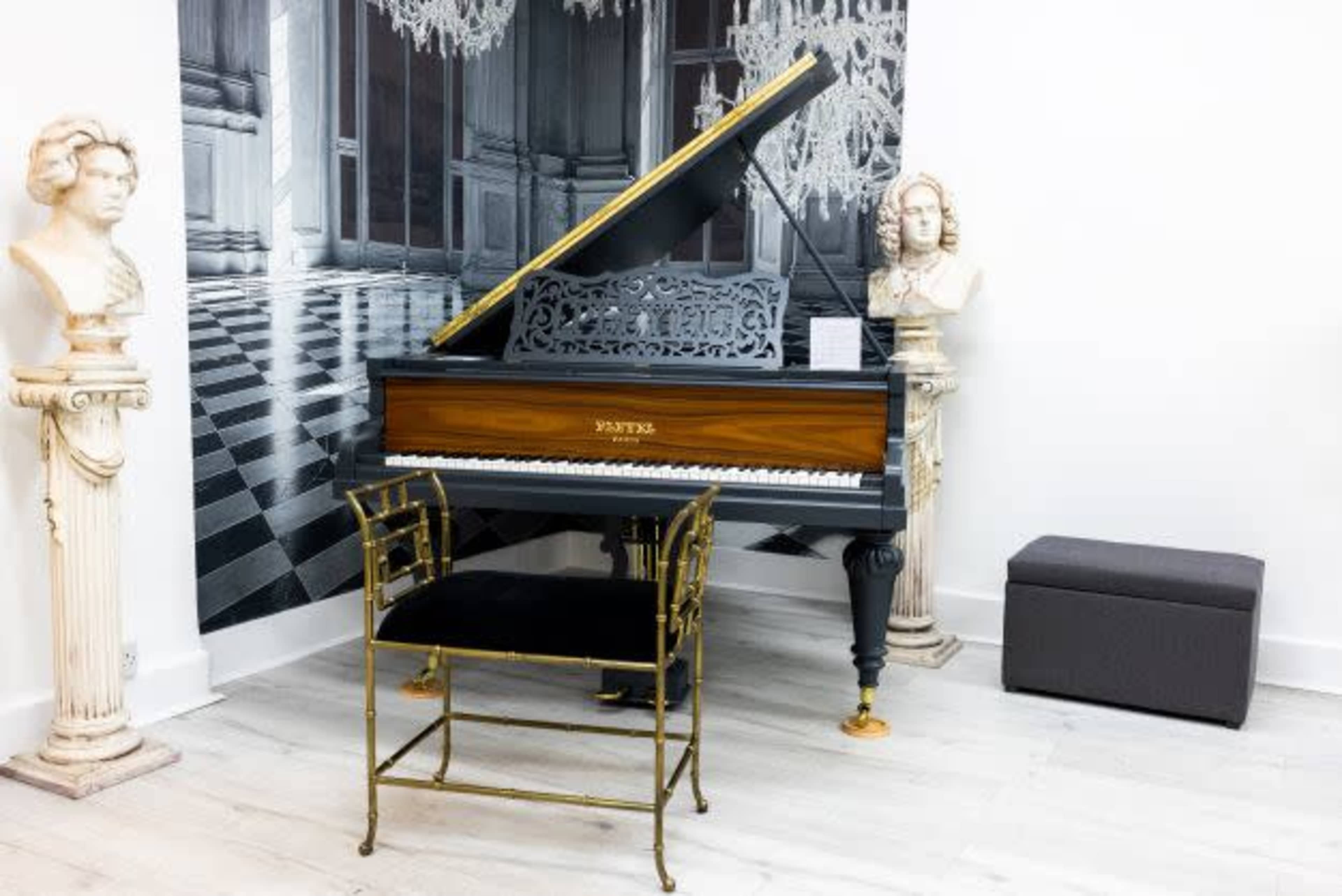 A grand piano with a black and gold finish, positioned in a room decorated with large black-and-white wall art and flanked by two classical busts on pedestals.