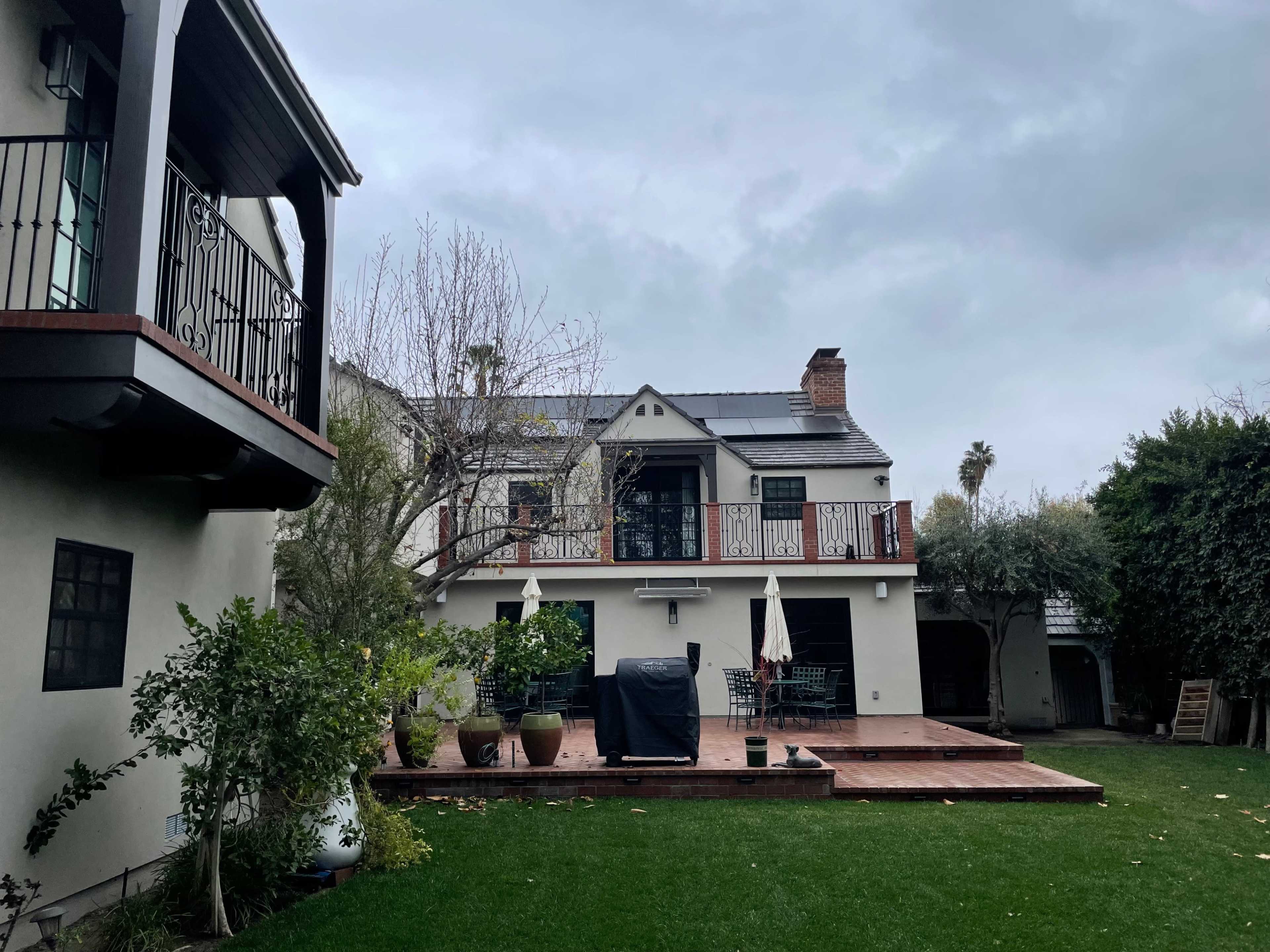 A backyard features a deck with a grill and seating area, surrounded by plants and greenery, with a two-story house in the background.