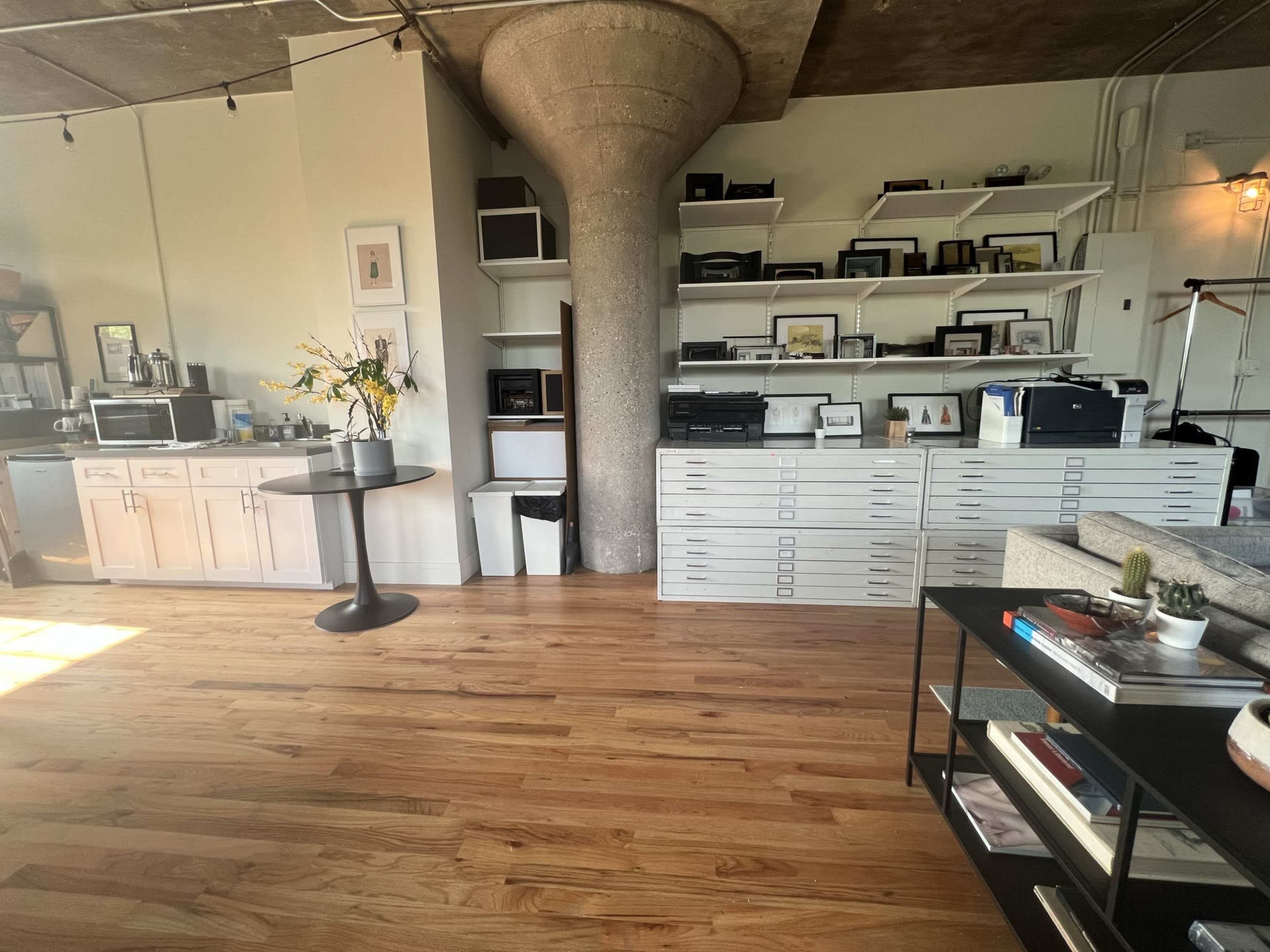 A modern office space featuring a round table, storage cabinets, printers, and shelves filled with framed photographs and office supplies, all set against a concrete pillar.