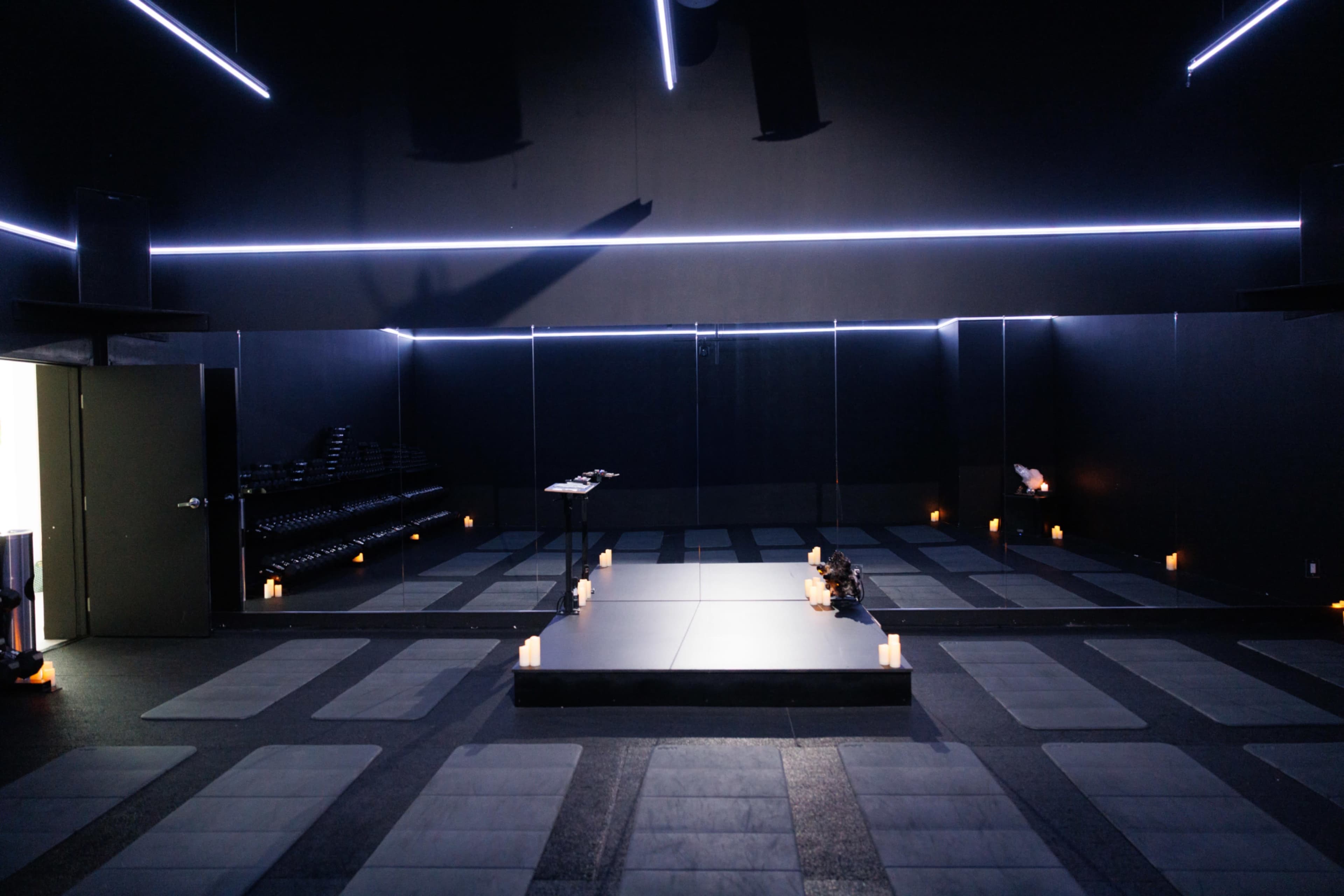 The image shows a dimly lit studio with a mirrored wall, soft flooring mats, and candles arranged on the ground, creating a tranquil environment for movement or meditation.