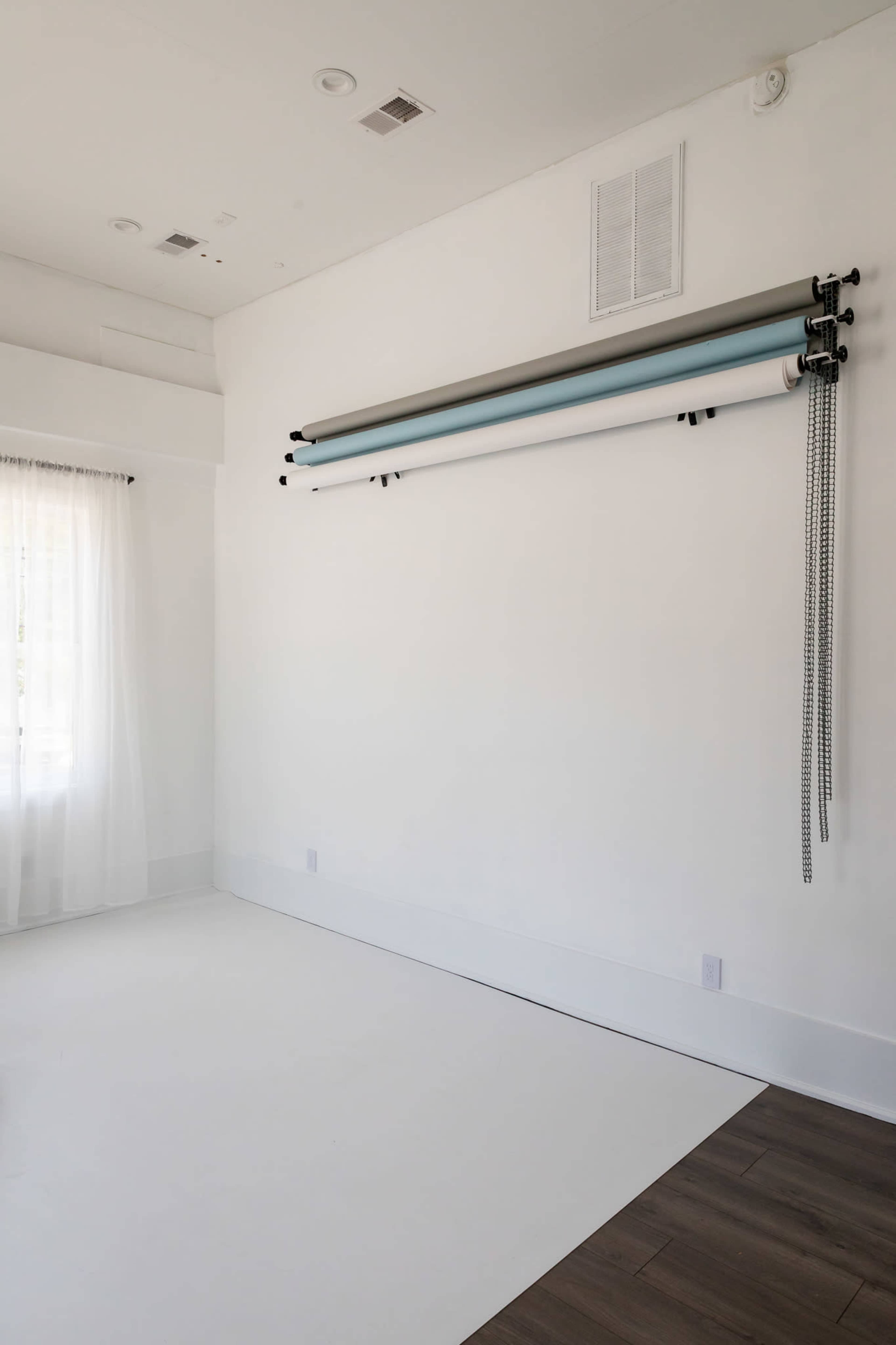 The image shows a minimalist studio space featuring a white wall, a large window with sheer curtains, and three rolls of backdrop paper mounted above the floor.