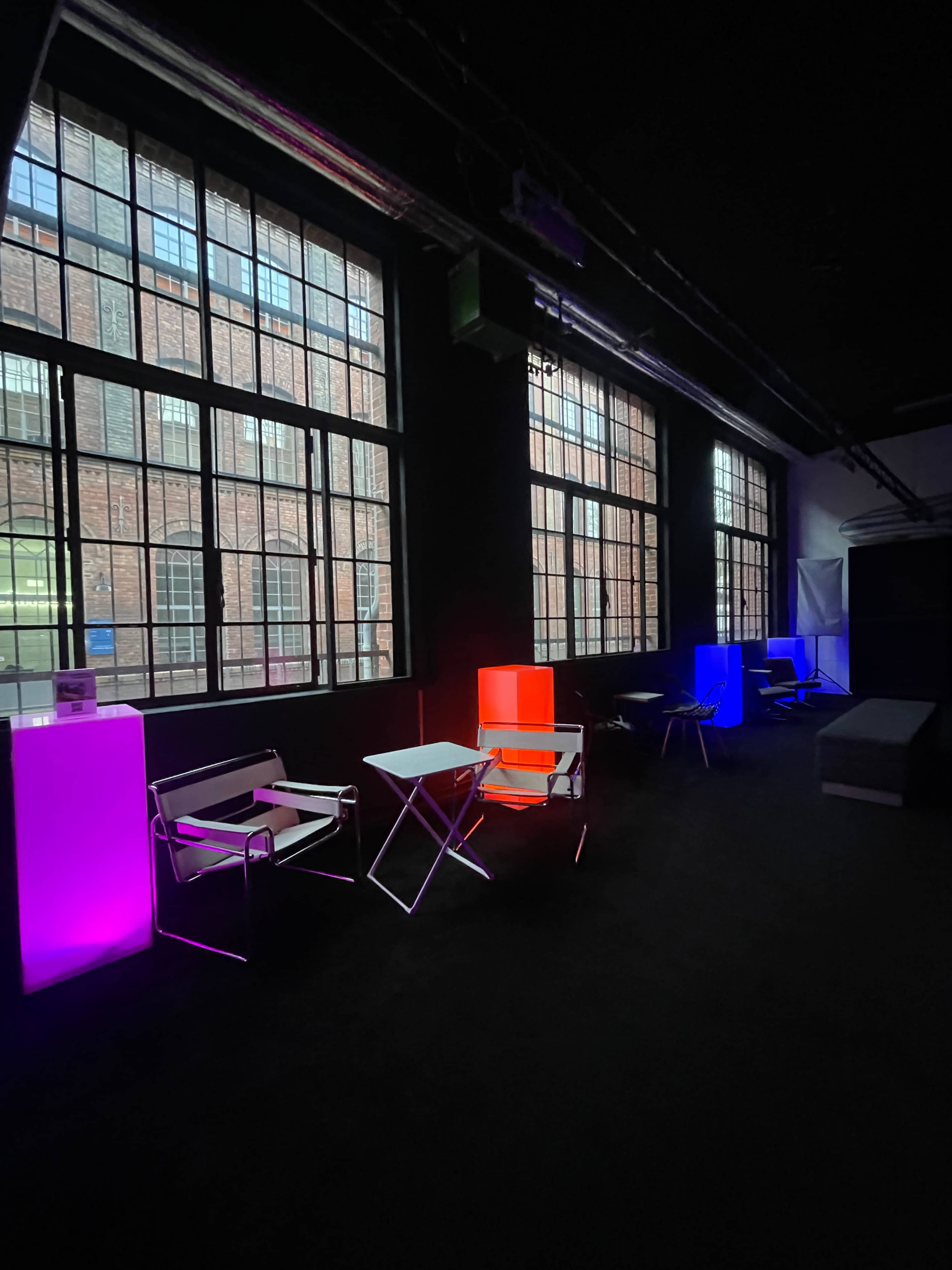 The image shows a dimly lit room with glowing furniture and large windows revealing a brick building outside.