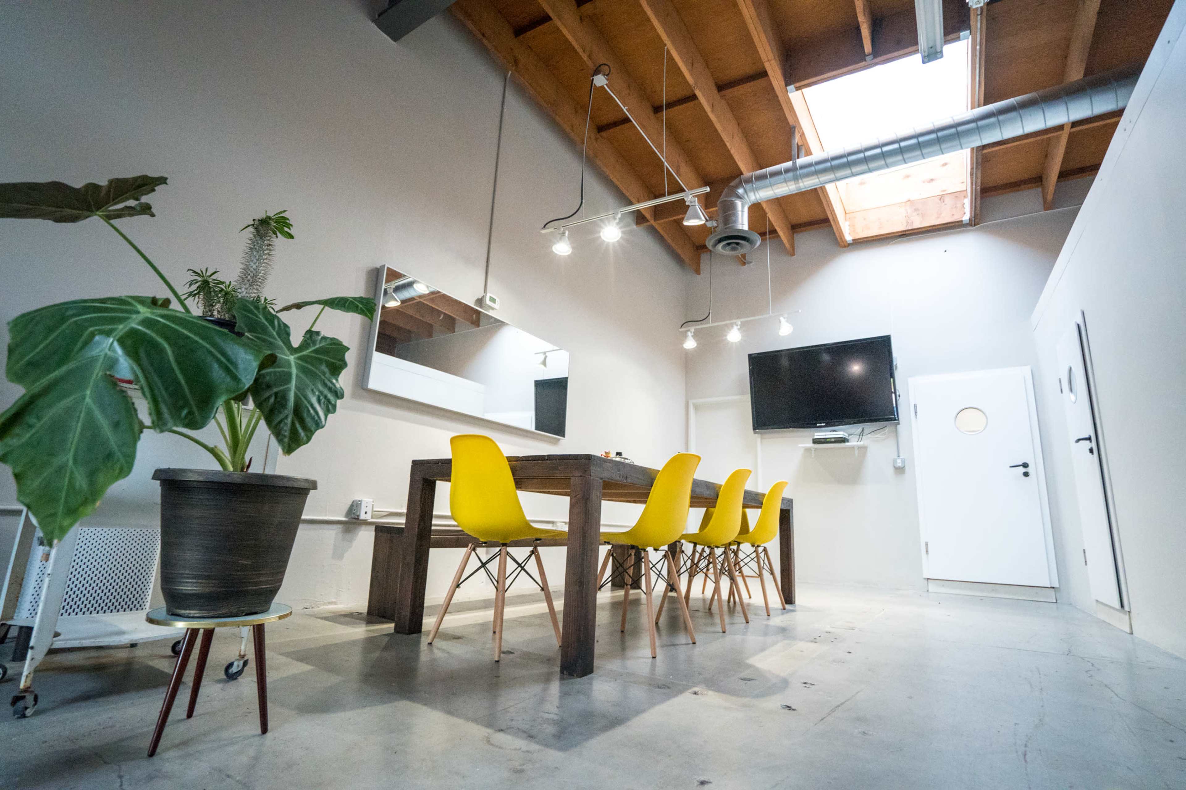 A modern conference room features a long wooden table surrounded by yellow chairs, large mirror, wall-mounted TV, and a skylight providing natural light.