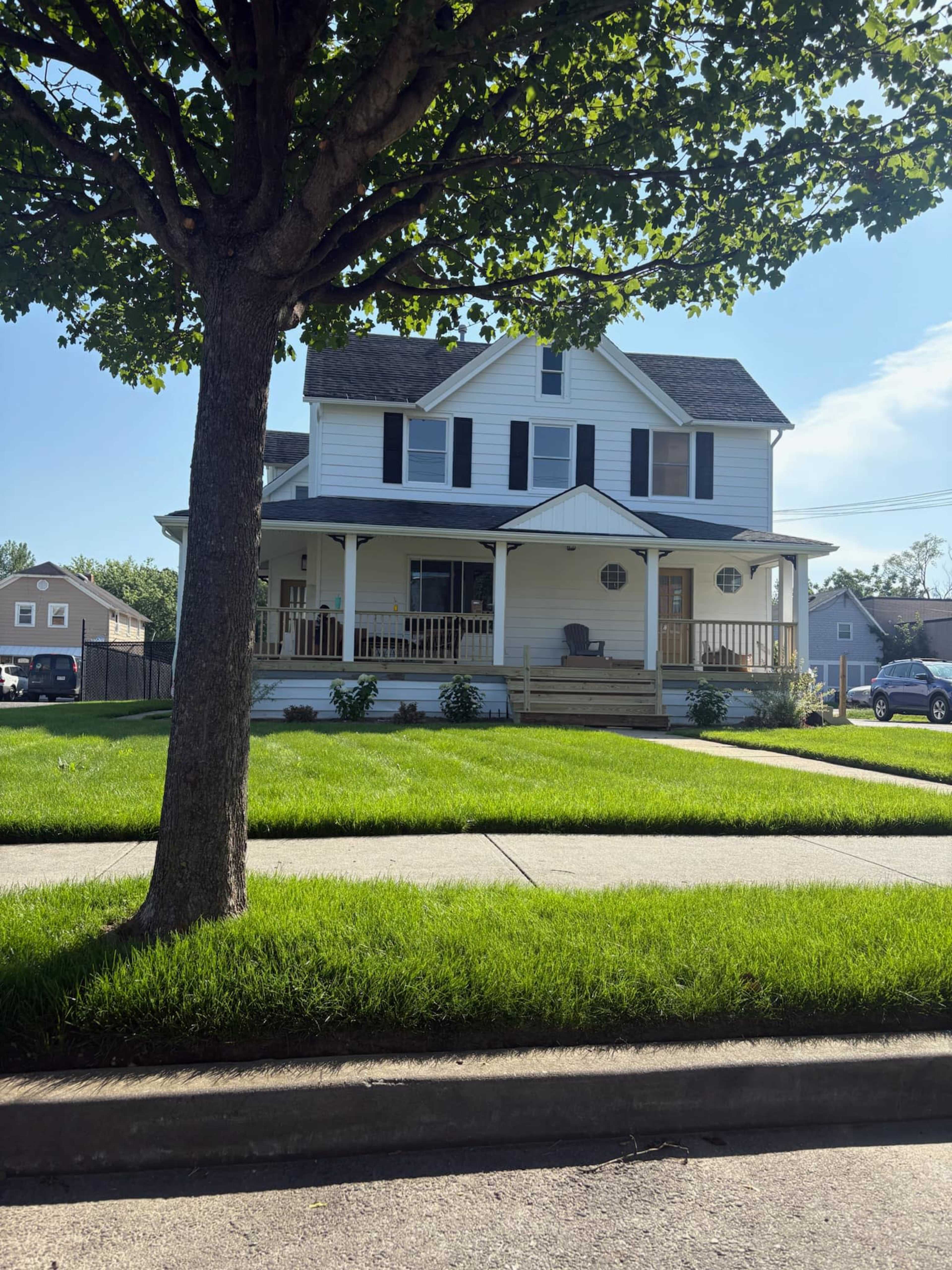 A two-story white house with a front porch and black shutters is surrounded by well-manicured grass and a tree.