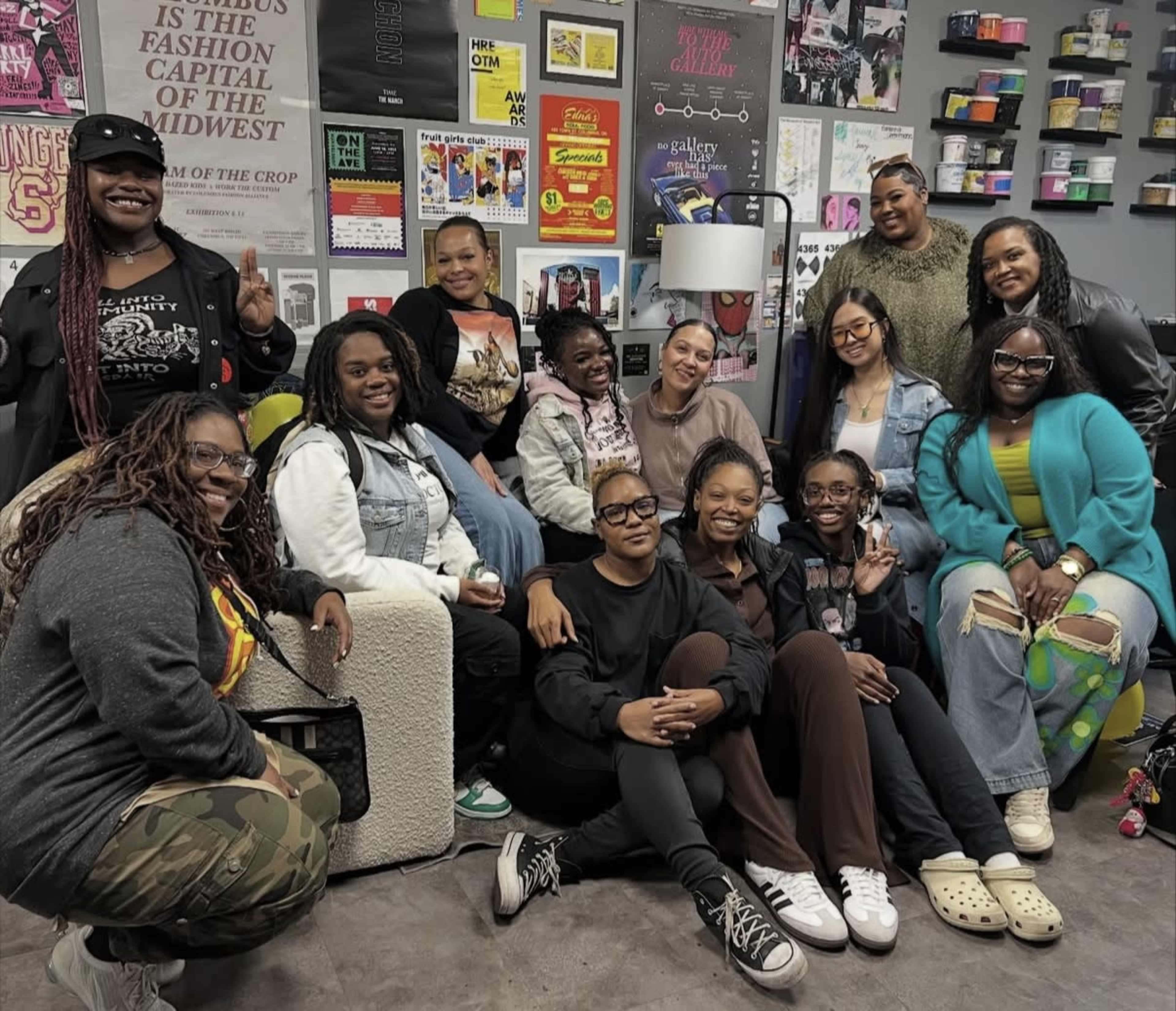 A diverse group of thirteen individuals poses together in a creative workspace filled with posters and colorful decor.