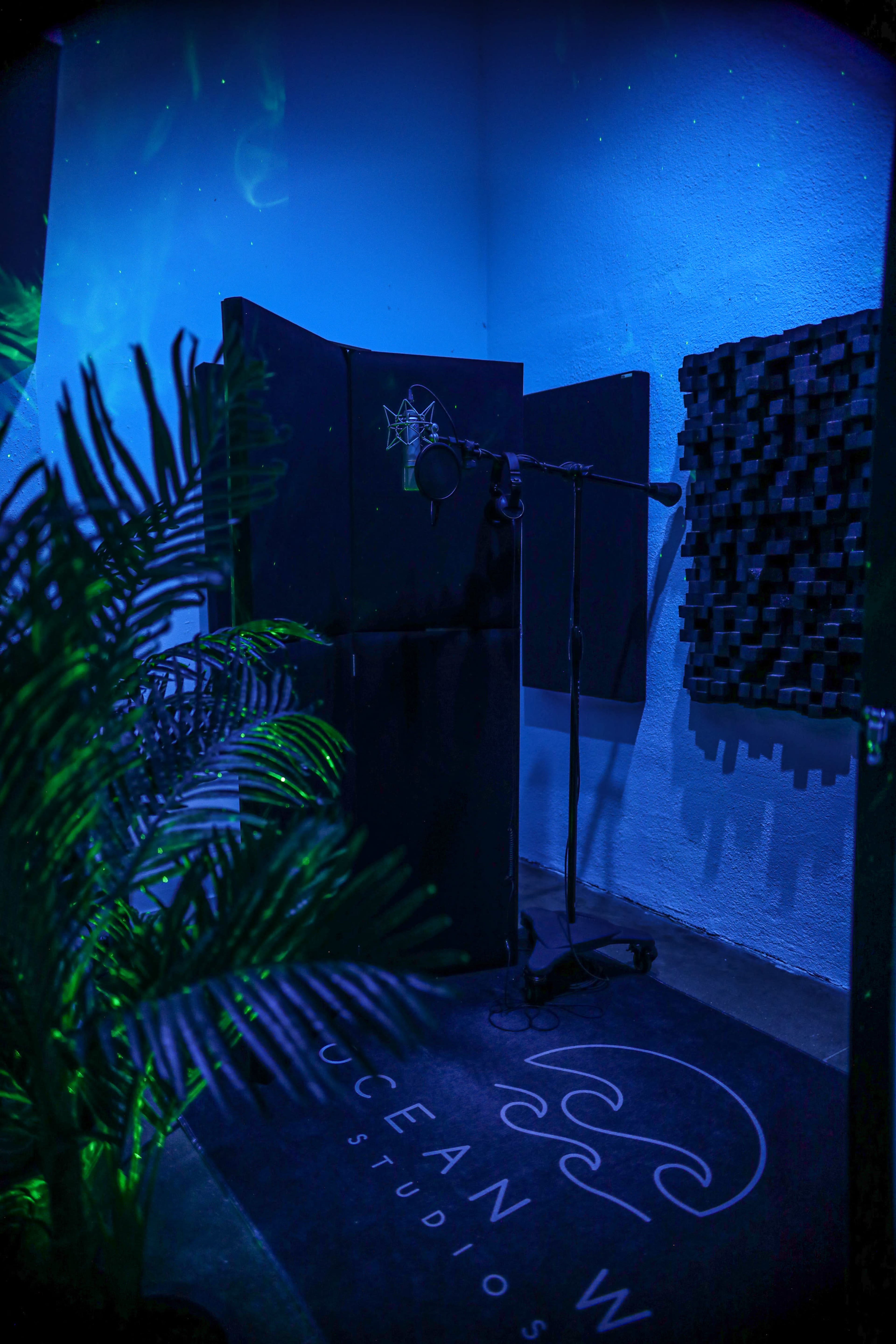 A recording studio corner is illuminated with blue lighting, featuring a microphone on a stand, acoustic panels, and a decorative rug.