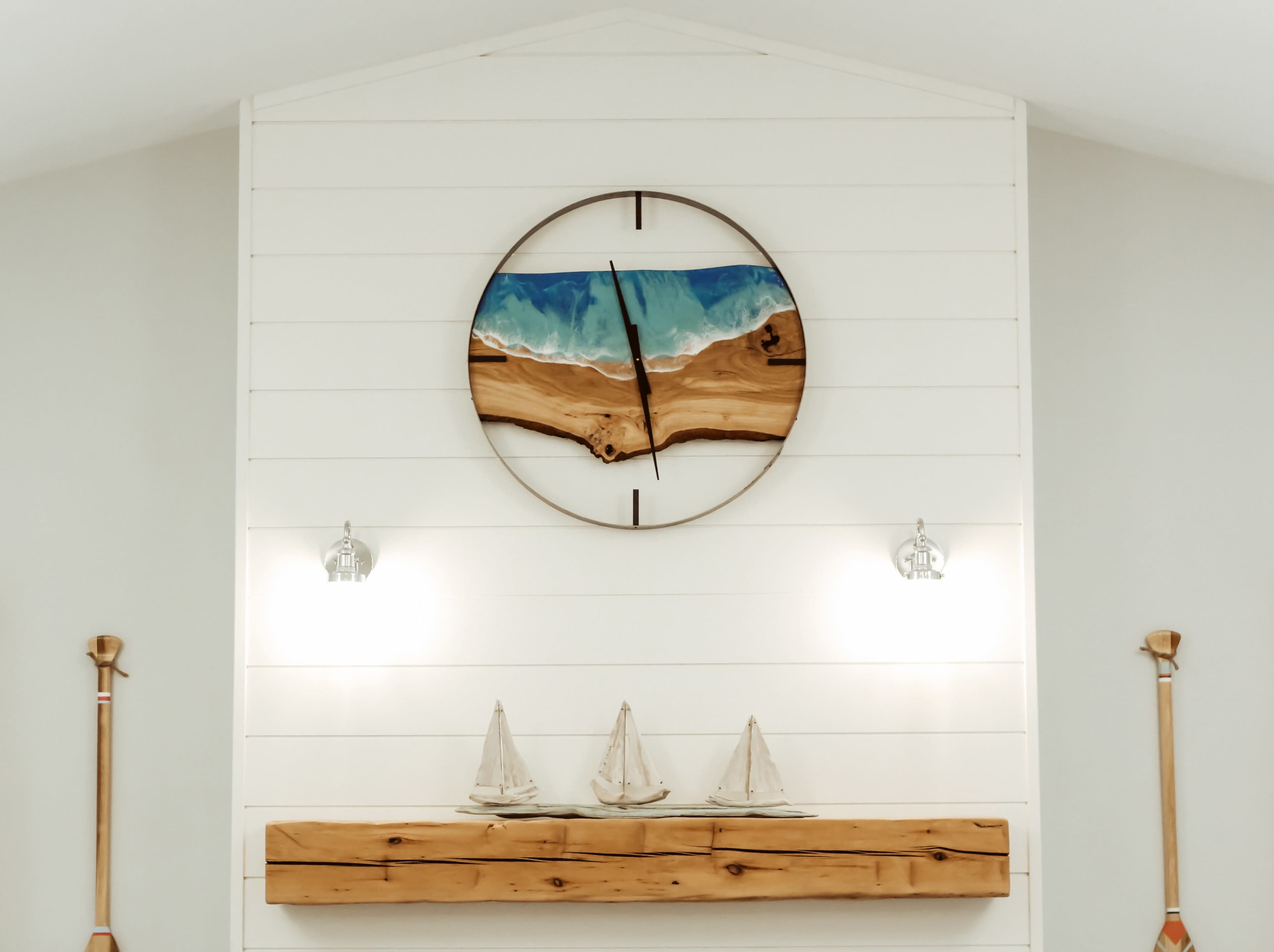 A wooden clock featuring a blue and tan design is mounted on a white shiplap wall above a shelf that holds three miniature sailboats.