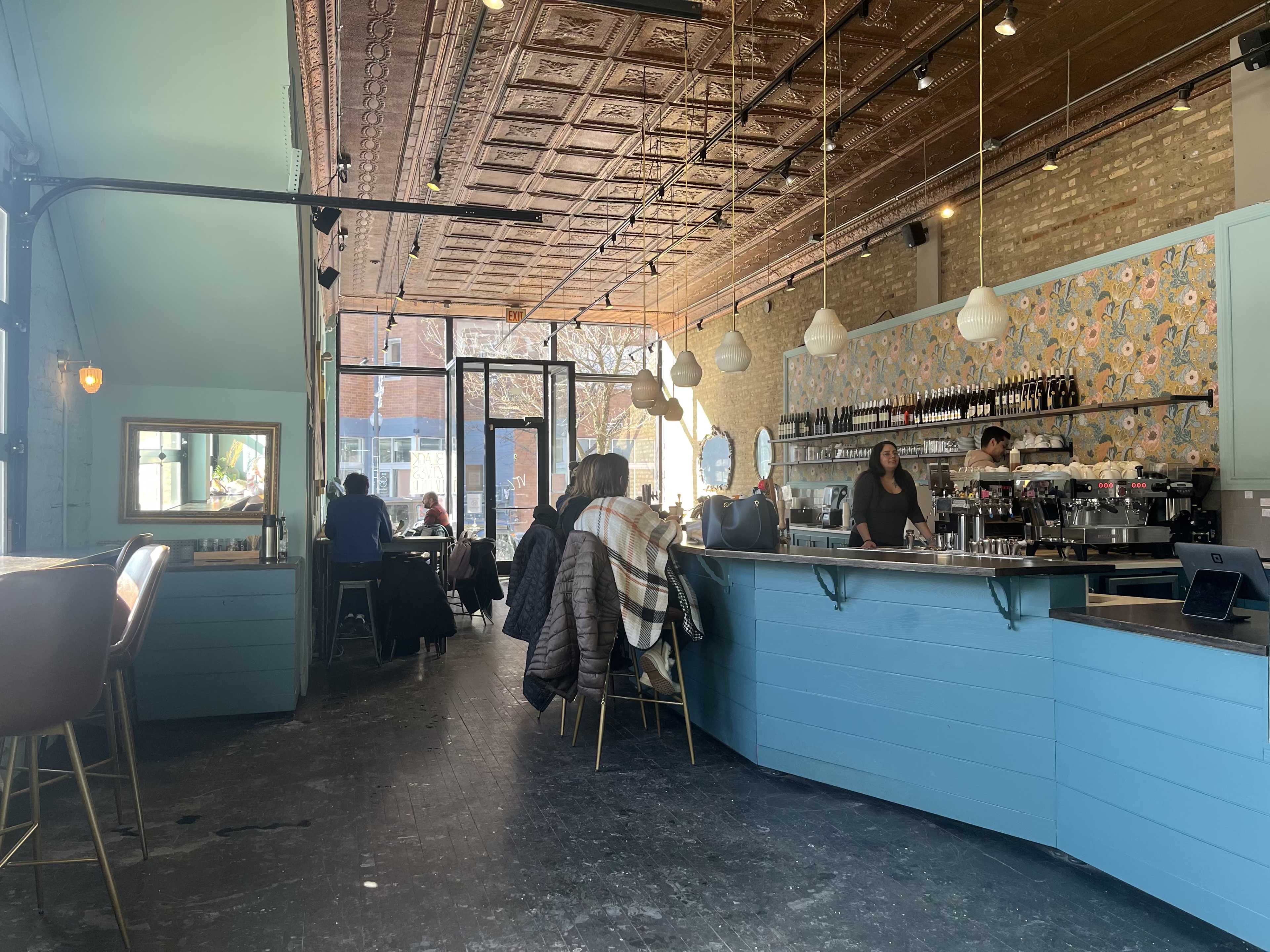 The image shows a bright cafe interior featuring a blue bar counter and patrons seated at tables, with a woman serving coffee in the background.
