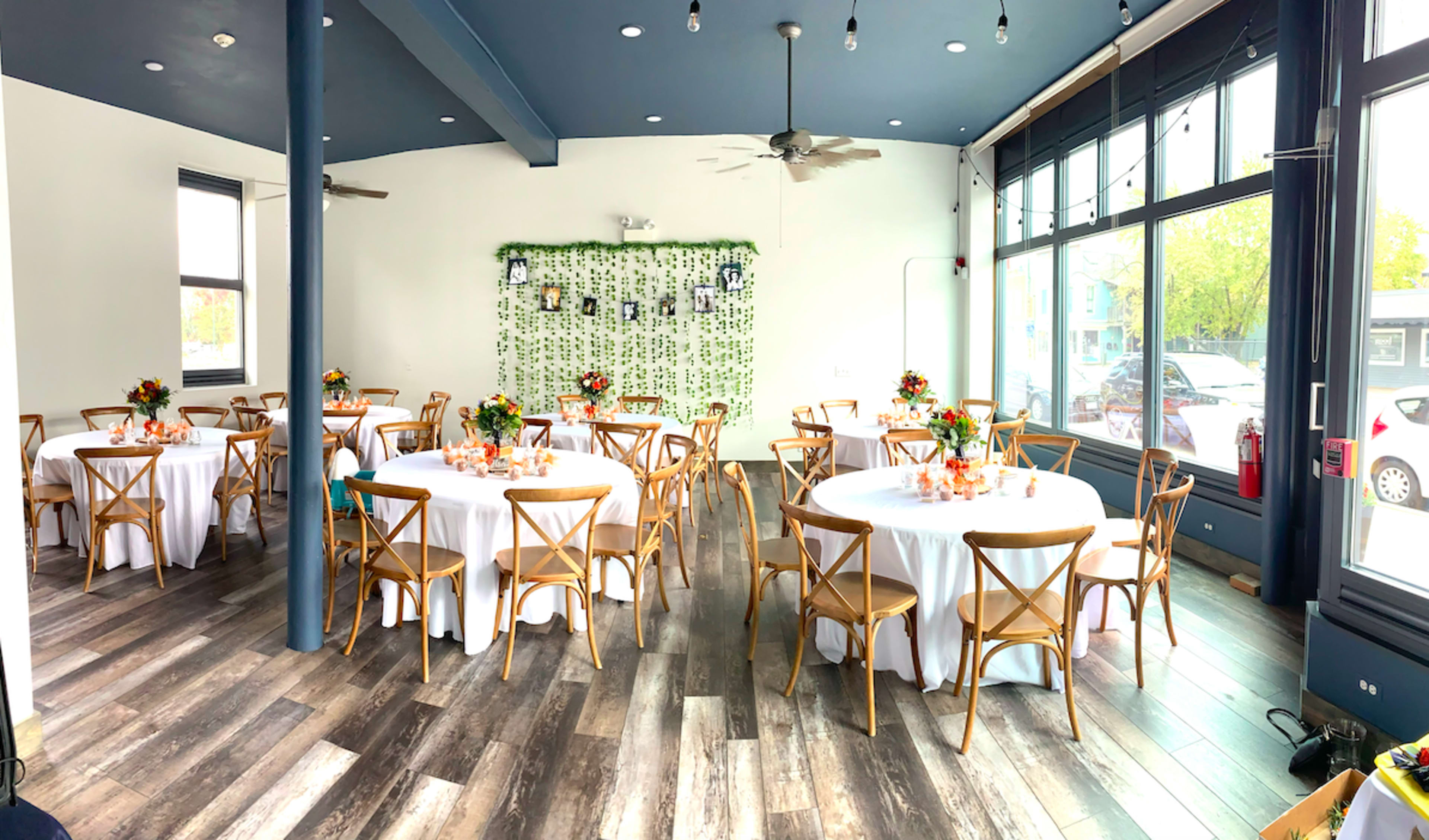 The image shows a decorated event space with round tables, white tablecloths, and centerpieces, surrounded by large windows and a greenery backdrop.