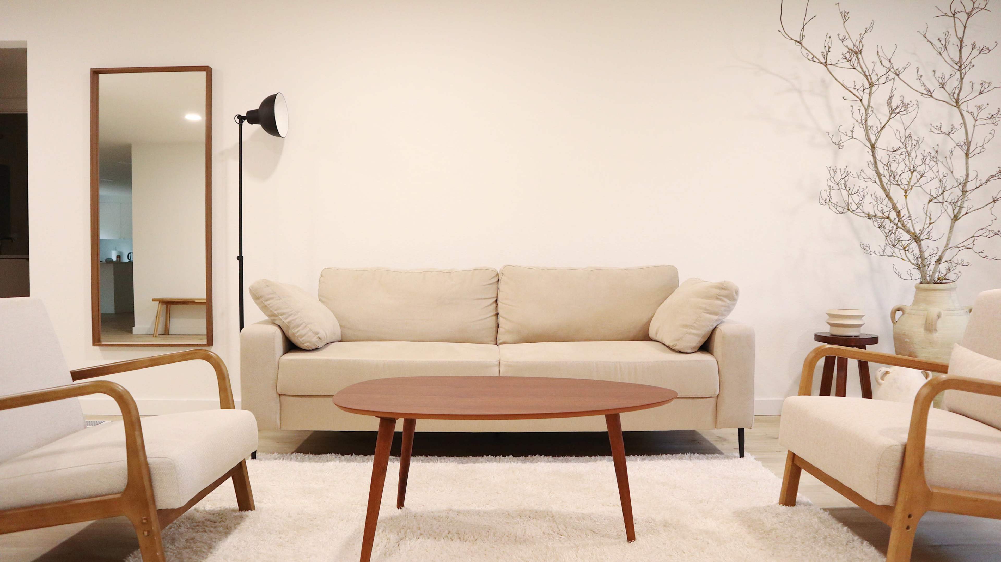 A modern living room with a beige sofa, two armchairs, a round wooden coffee table, and a tall mirror against a white wall.