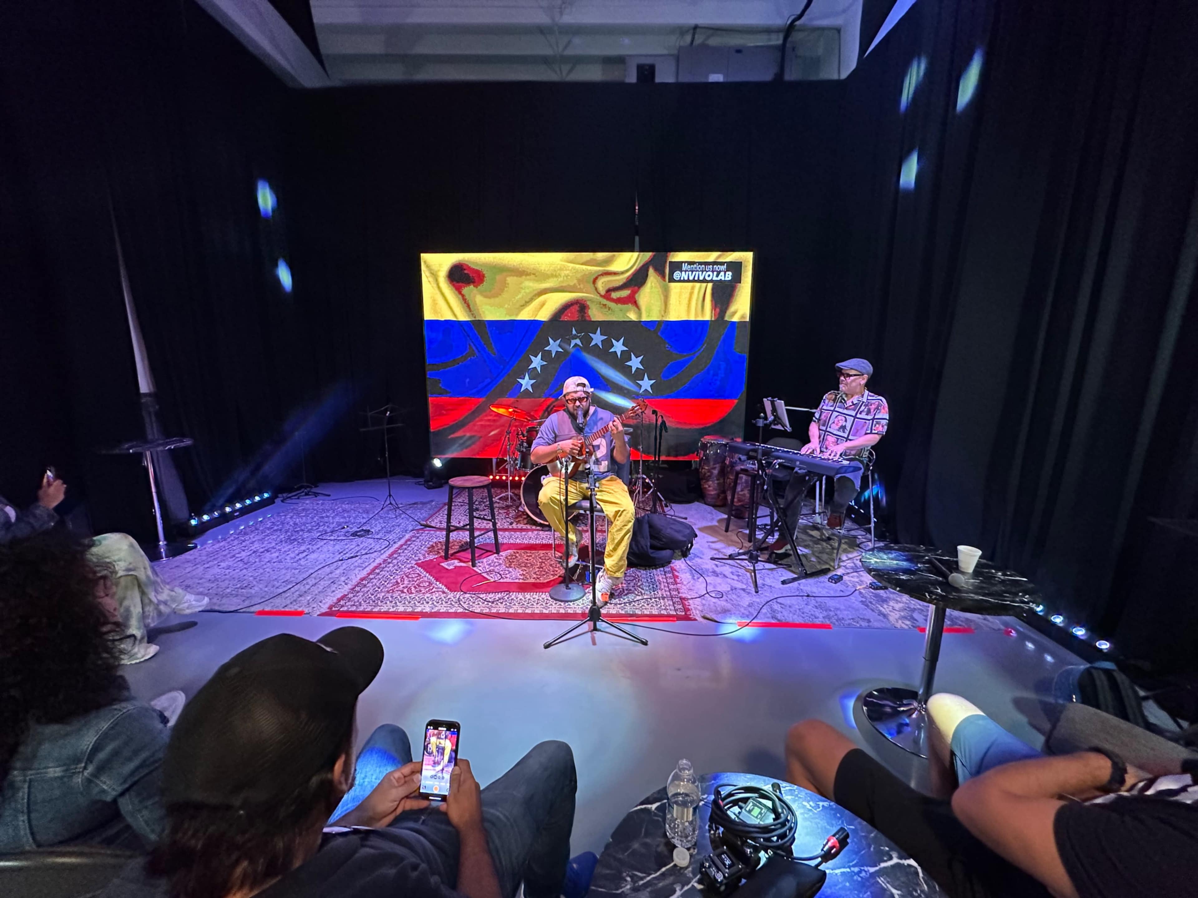 A musician performs on stage in front of a Venezuelan flag backdrop while a second musician plays the keyboard nearby.