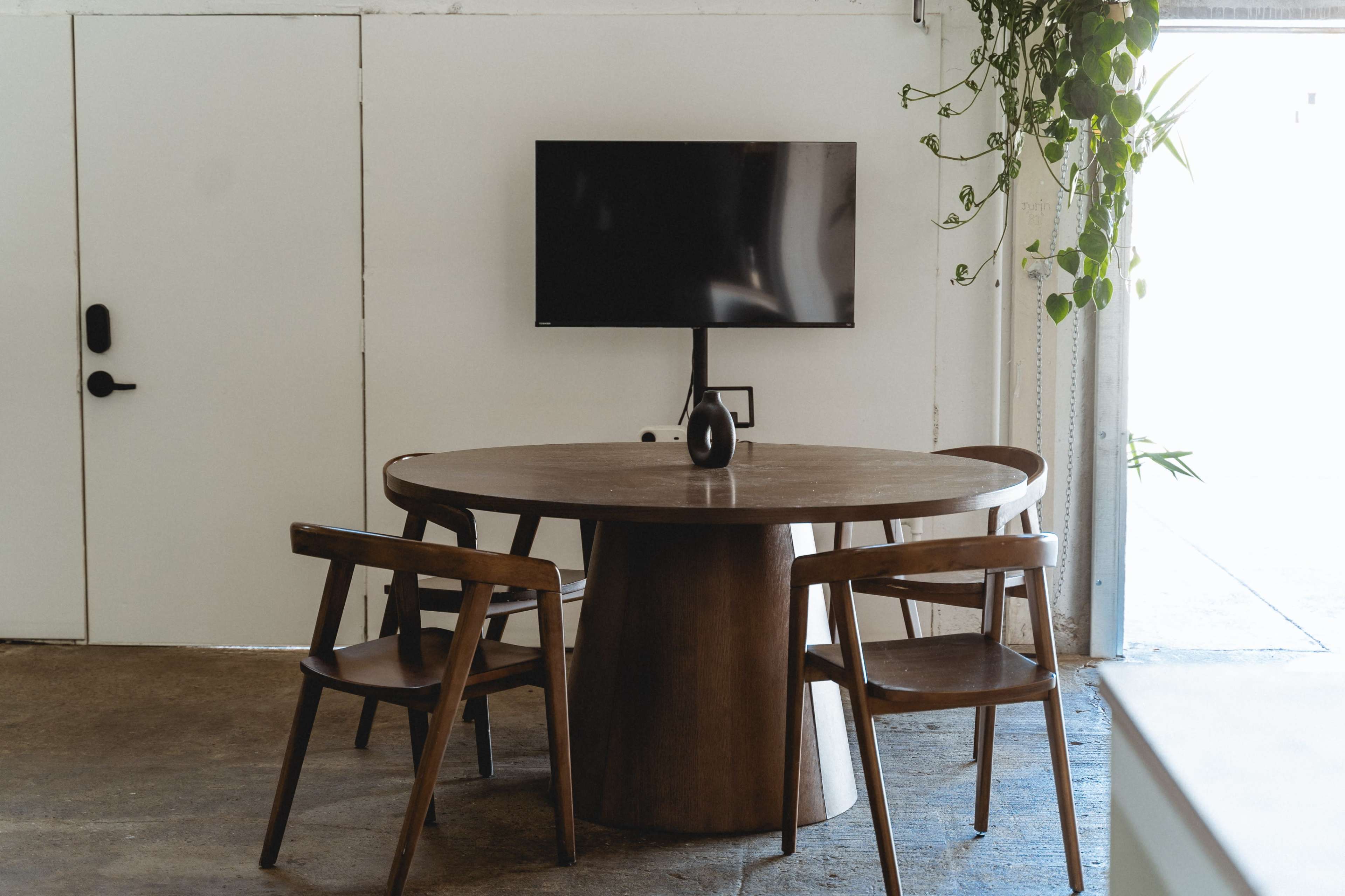 A round wooden table with three chairs is positioned in a room featuring a mounted television and a potted plant.