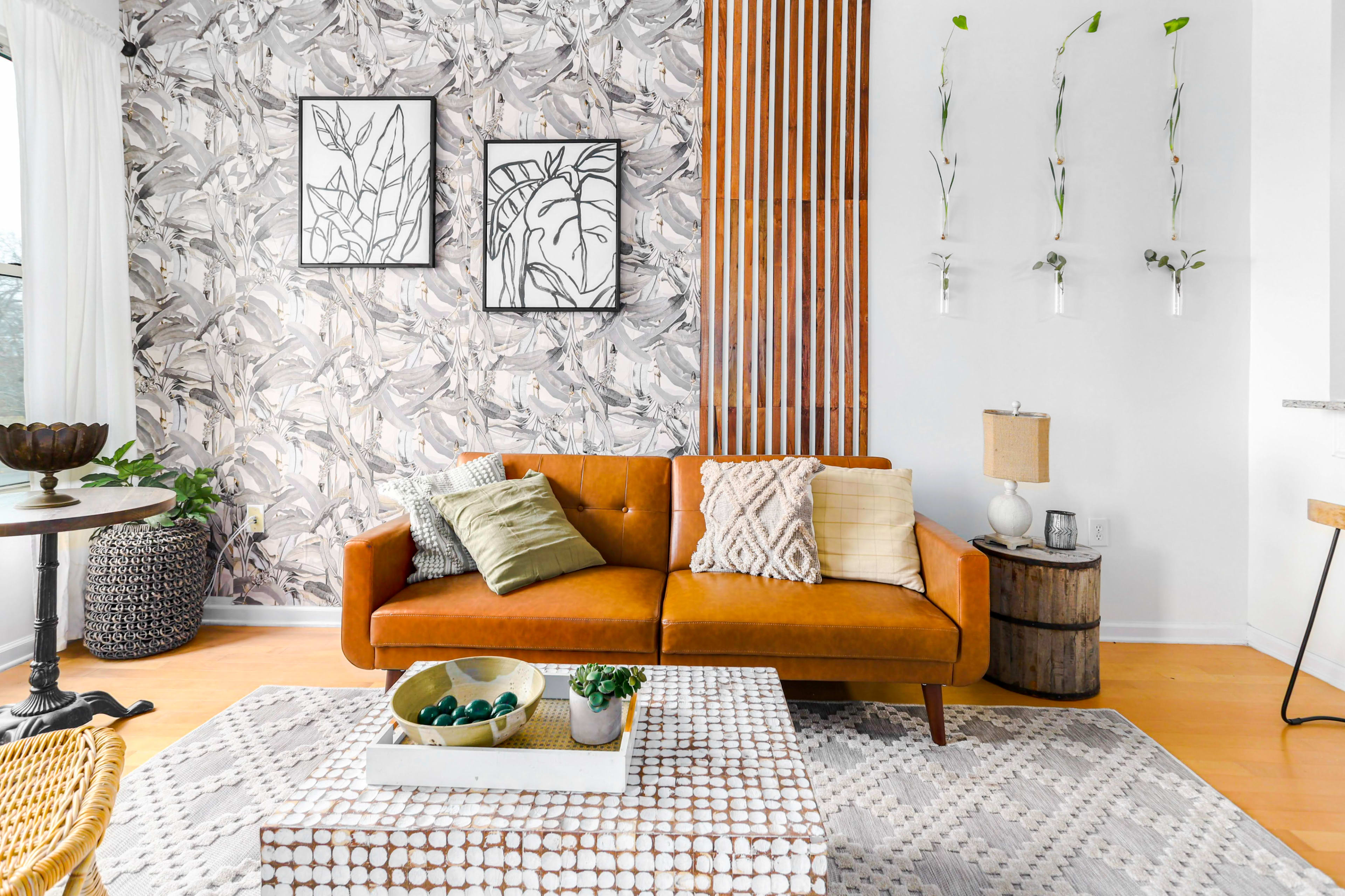 The image shows a stylish living room featuring a brown leather sofa, patterned wallpaper, two framed artworks, a coffee table with decorative items, and a wooden side table.