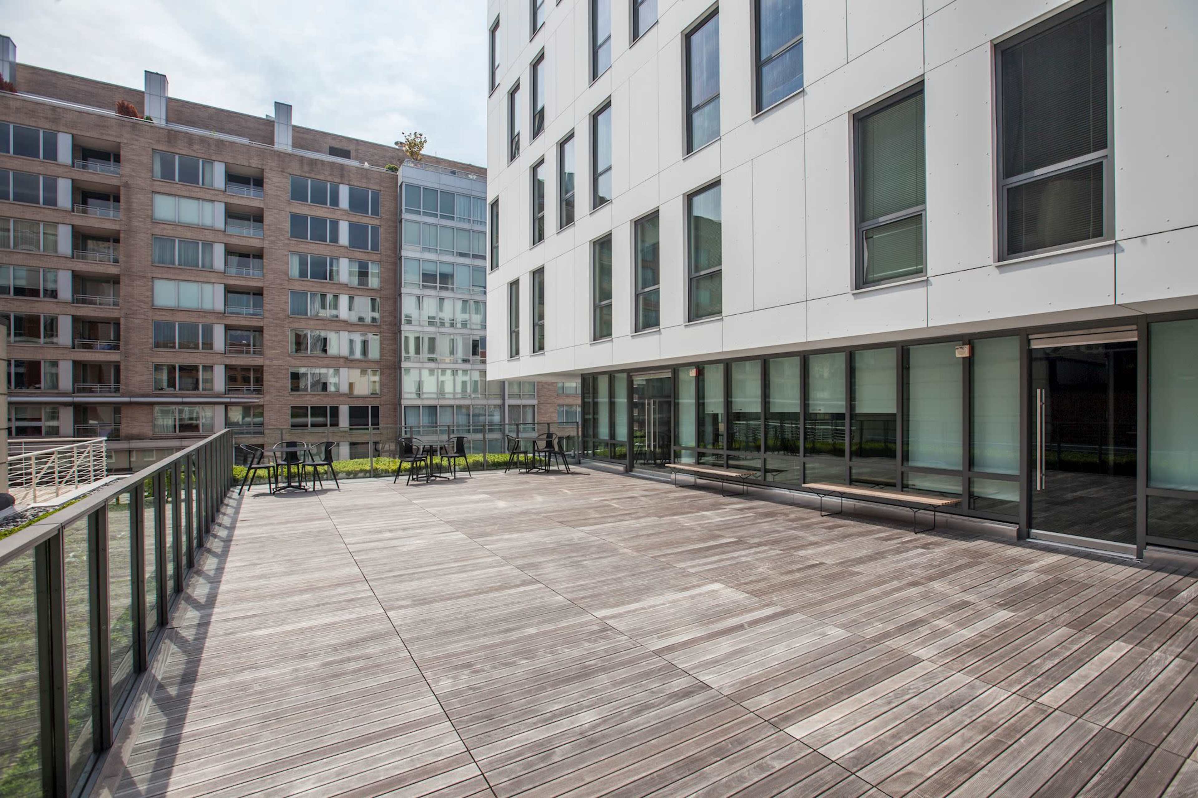 The image shows a modern outdoor terrace with wooden flooring adjacent to a building with large glass windows.