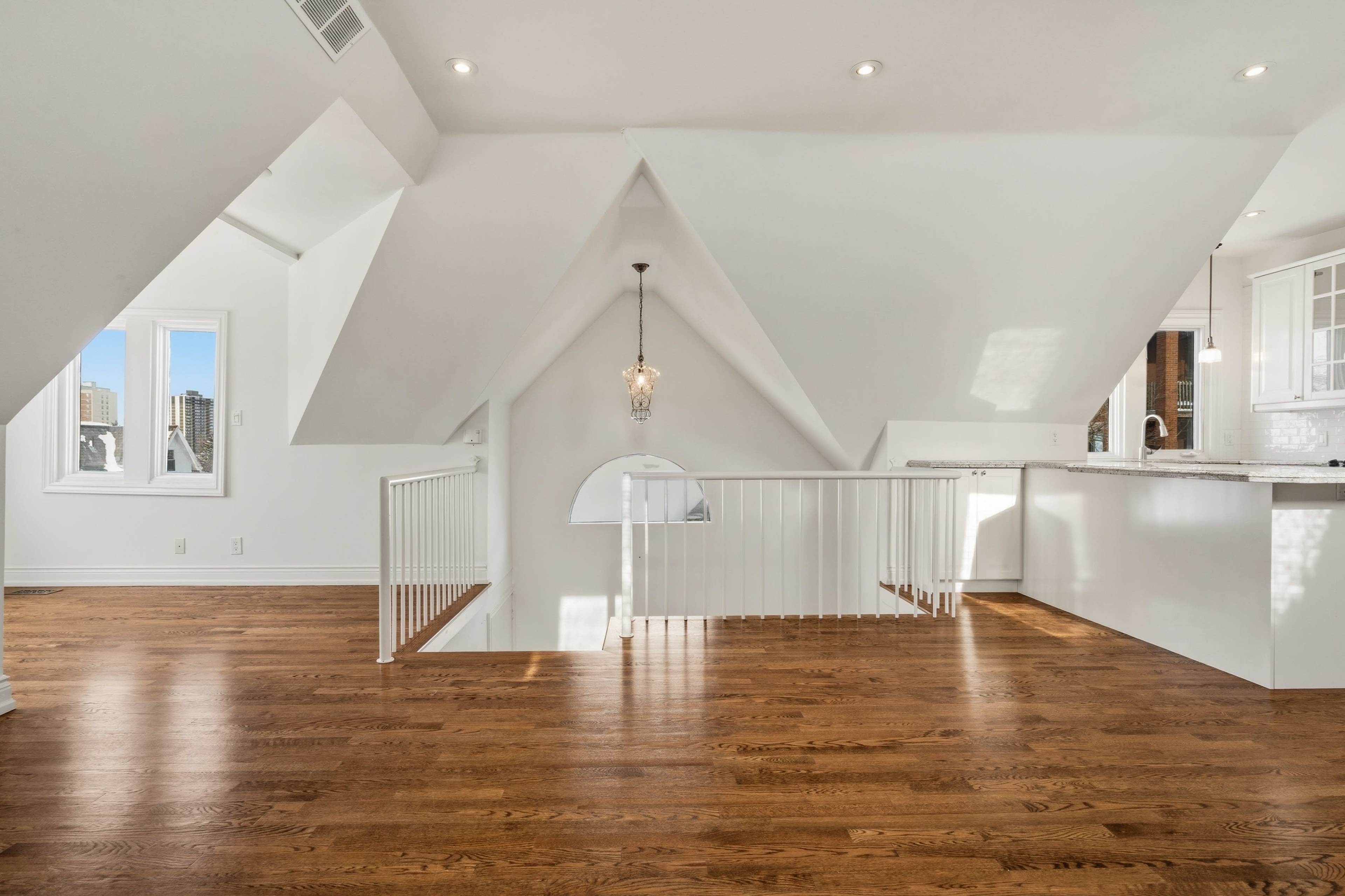 The image shows a bright, airy loft space with slanted ceilings, hardwood floors, and a modern railing leading to a kitchen area.